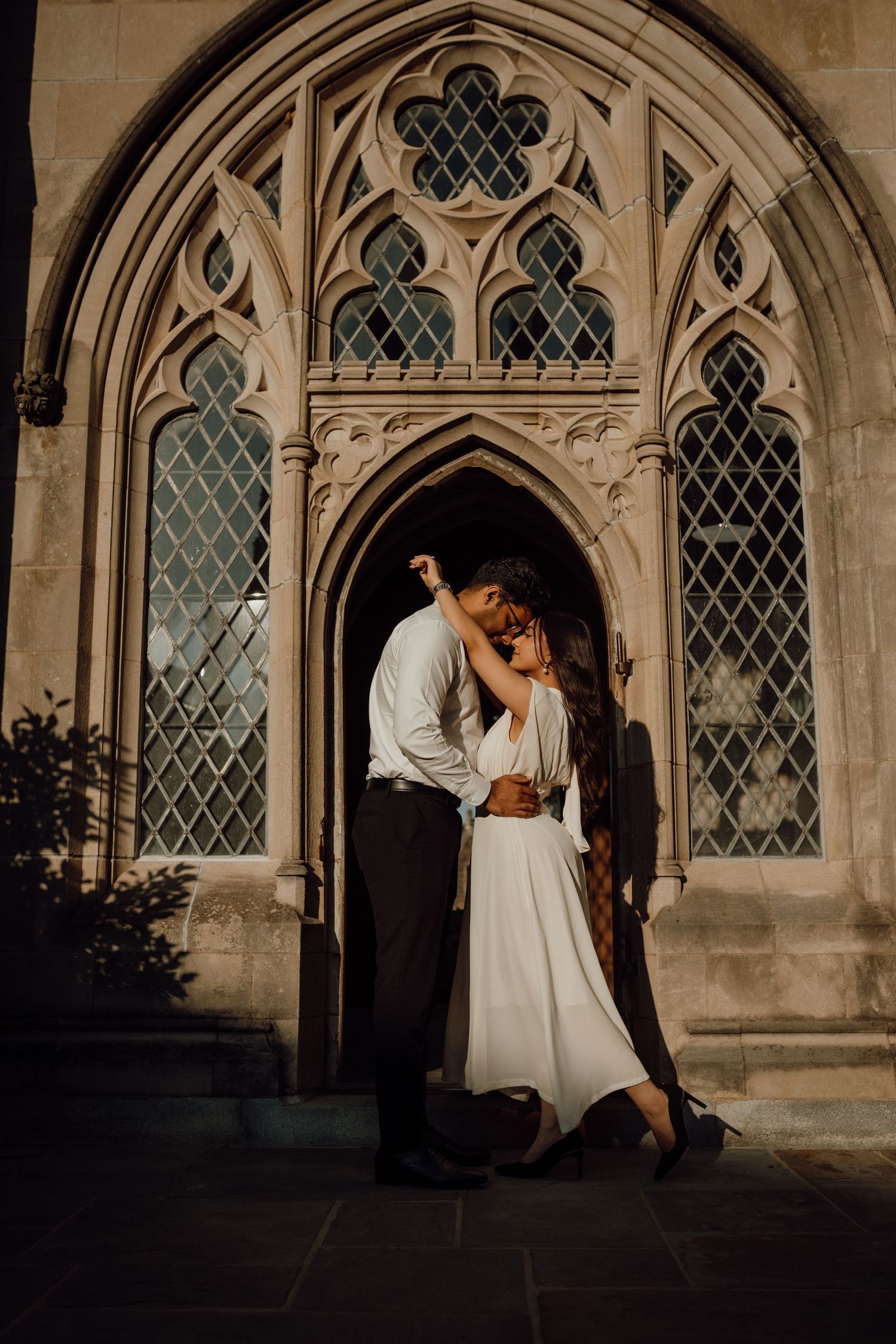 Couple kiss in front of an ornate Gothic stone doorway with diamond-pane windows in warm golden light.