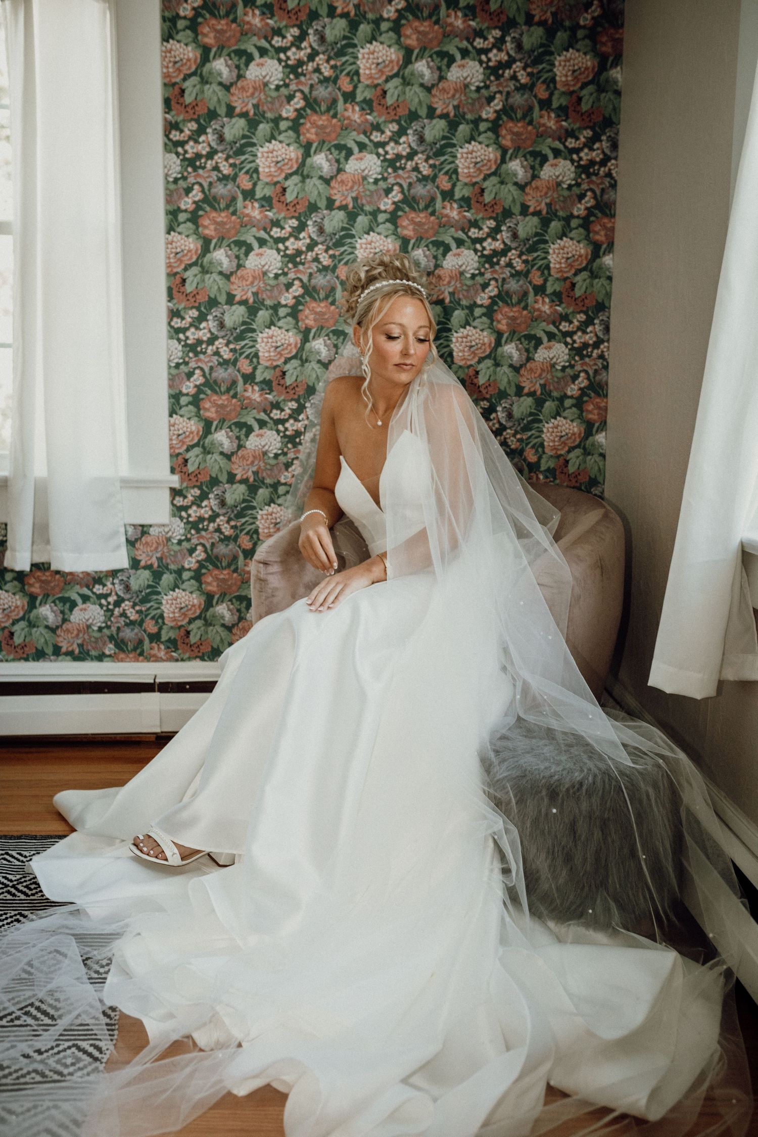 Bride in a flowing white gown and long veil sits by a window in front of floral wallpaper.