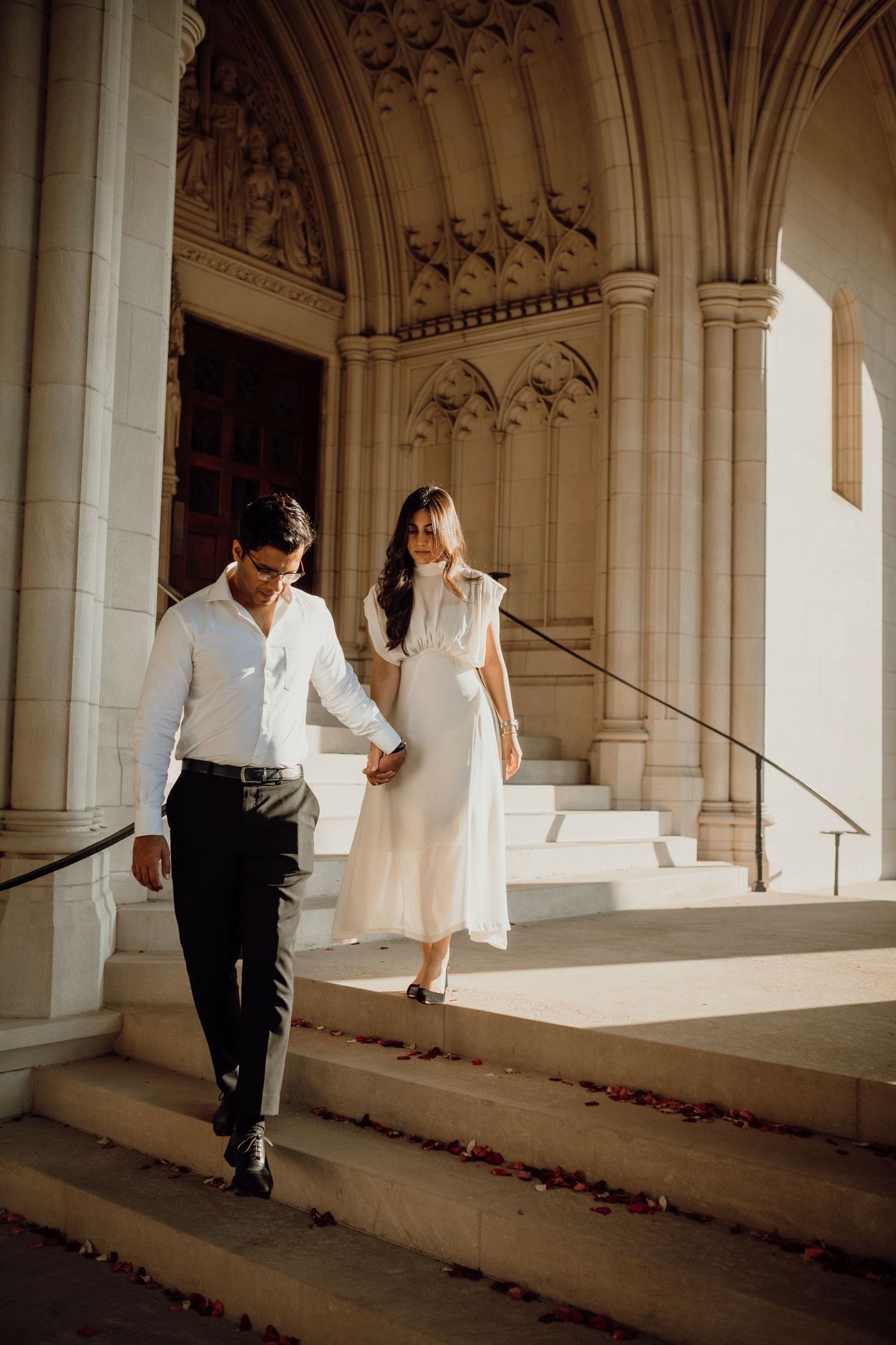 Couple holds hands while walking down the stone steps of a historic arched church entrance in golden light.
