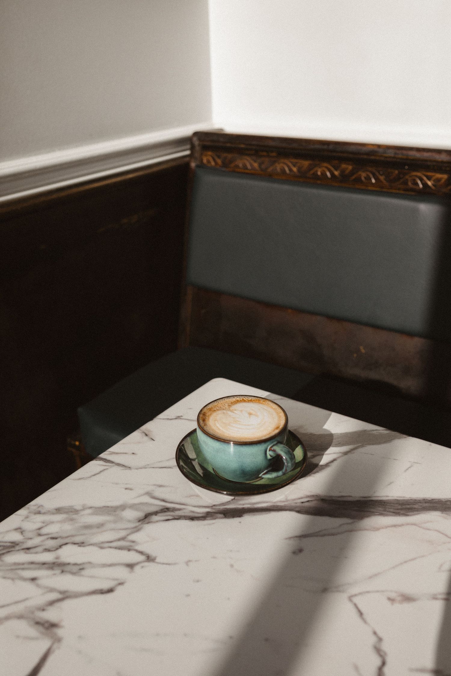 Latte with foam art in a teal ceramic cup on a marble café table beside a green leather booth.