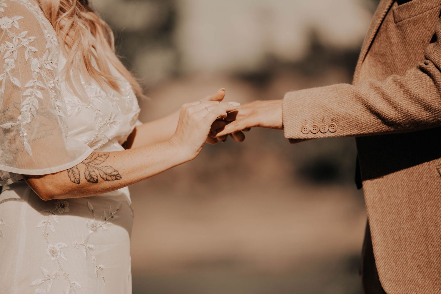 Bride in a lace wedding dress with a floral arm tattoo holds hands with a groom in a tweed jacket during a ring exchange.
