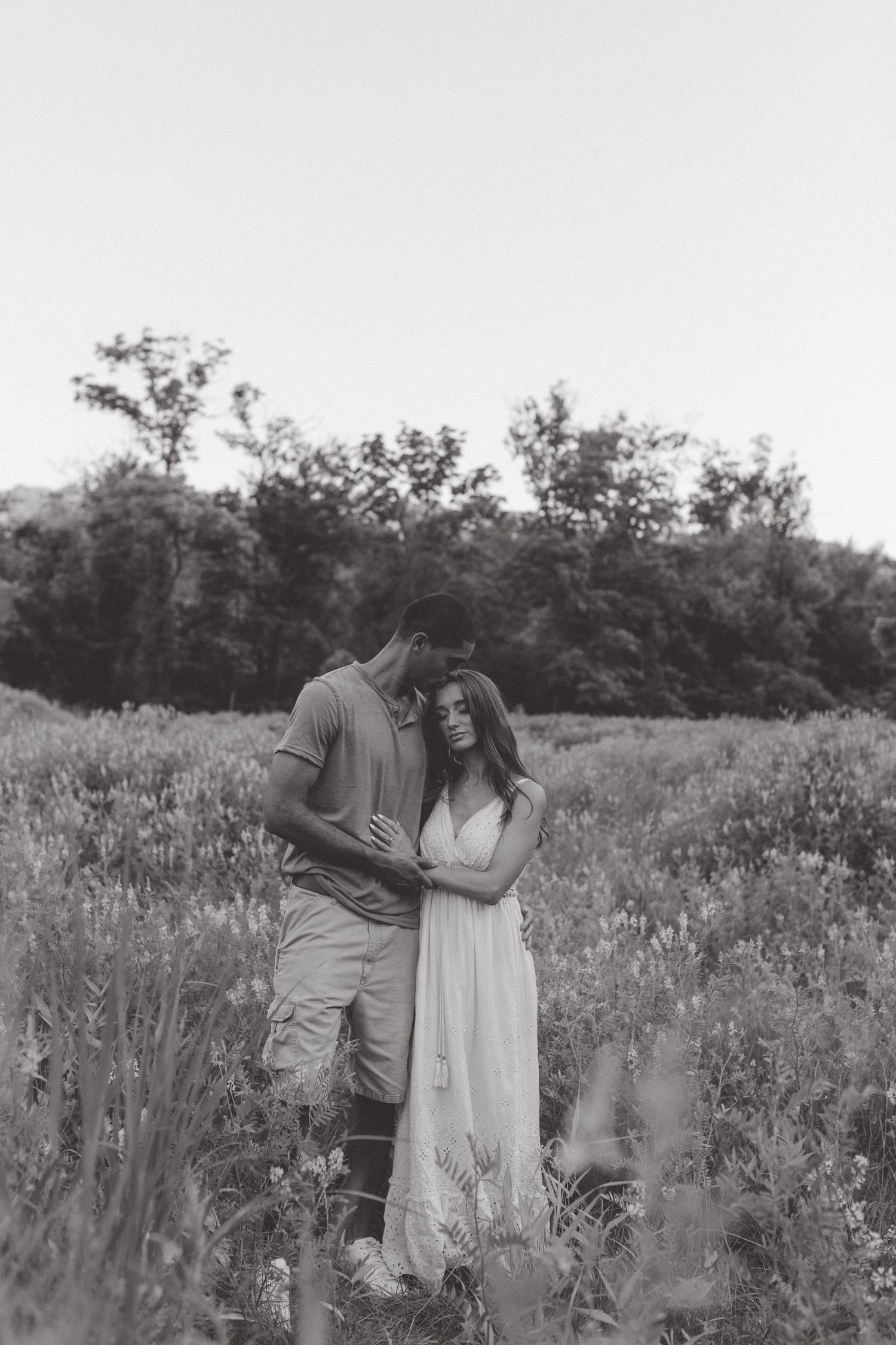 A black and white photo of a couple embracing in a wildflower meadow with trees in the background.
