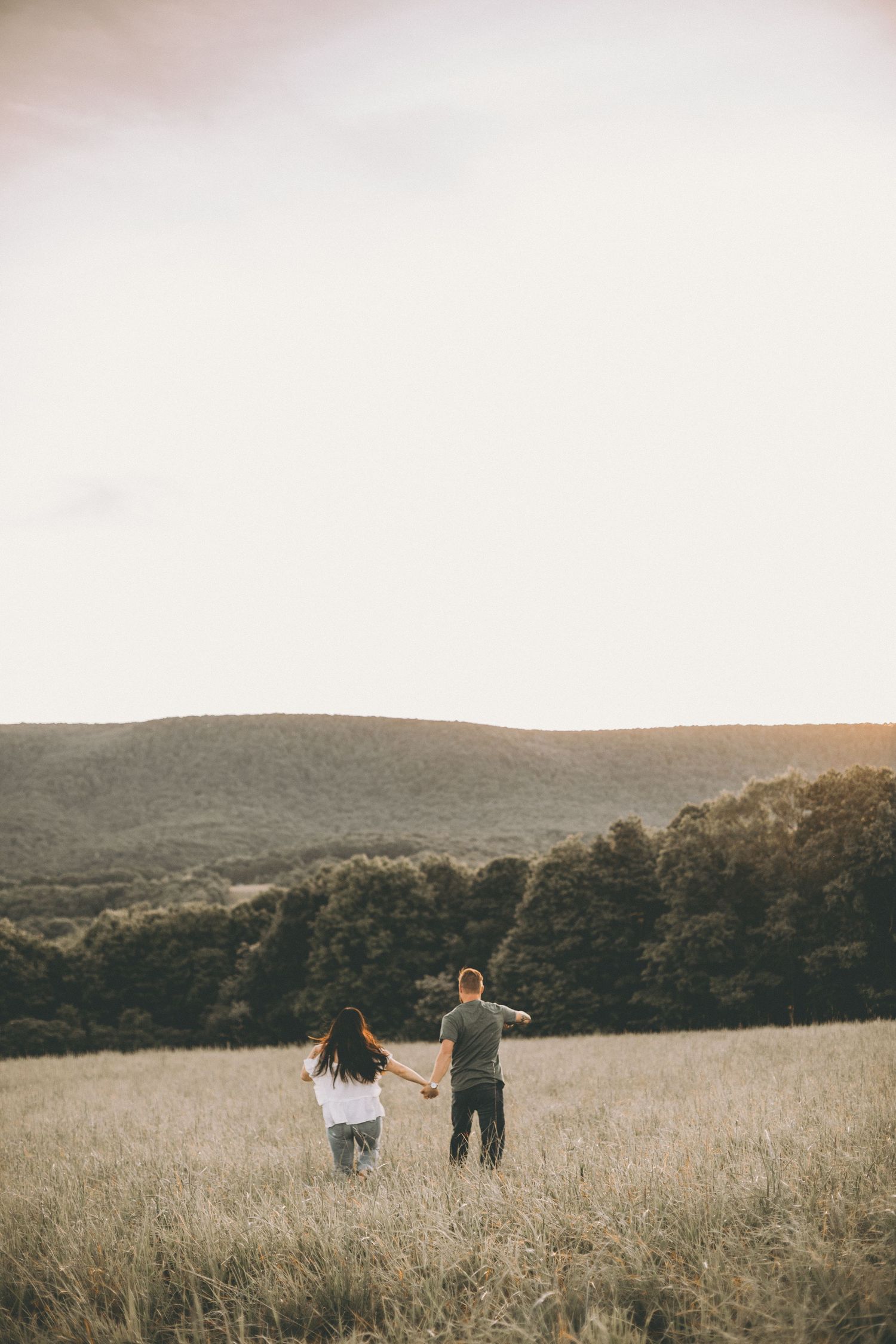 Couple holding hands and walking through a golden grass meadow toward forested hills under a soft pastel sky at sunset.