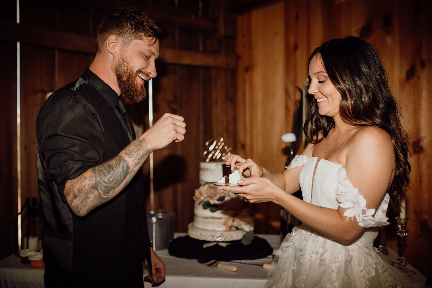 Smiling tattooed groom and bride in an off-shoulder lace gown feed each other wedding cake during a rustic barn reception.