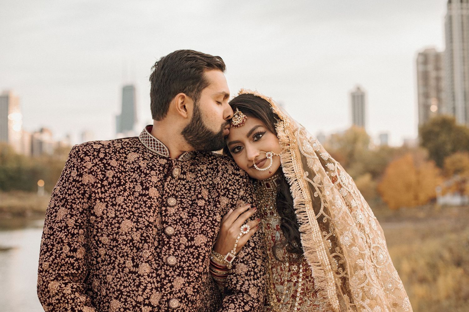 Bride and groom in embroidered traditional South Asian wedding attire embrace outdoors with a city skyline in the background.