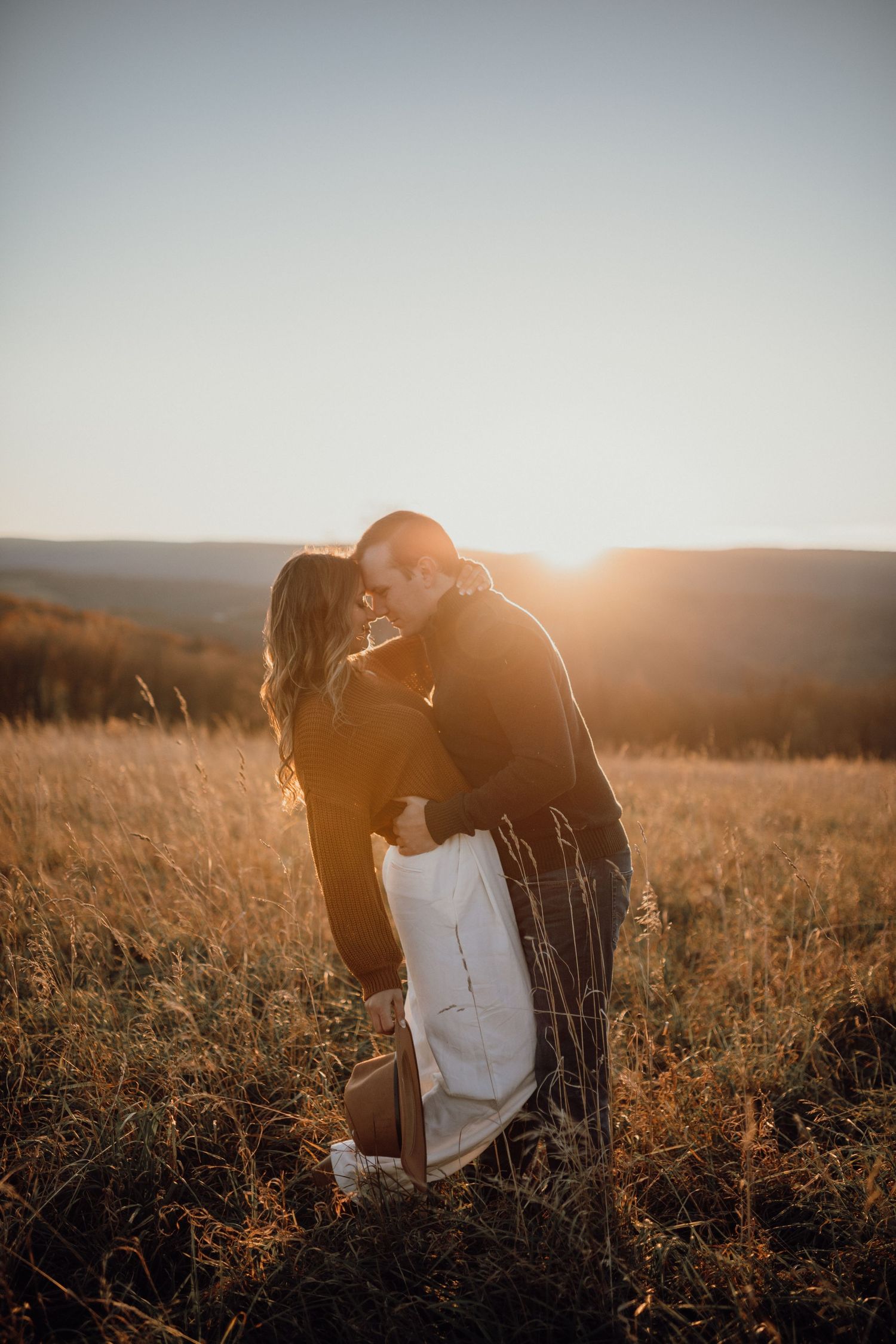 Couple touching foreheads in a golden field at sunset with a hat in hand and hills behind them.