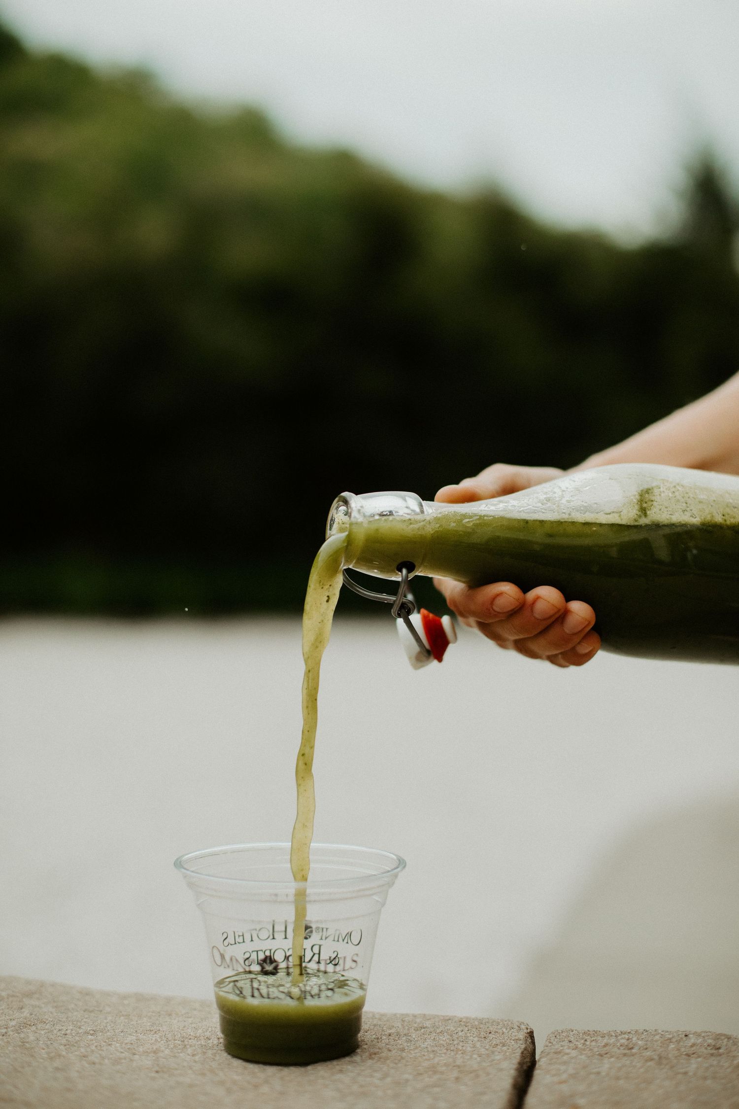 Hand pouring fresh green juice from a glass swing-top bottle into a clear cup outdoors.