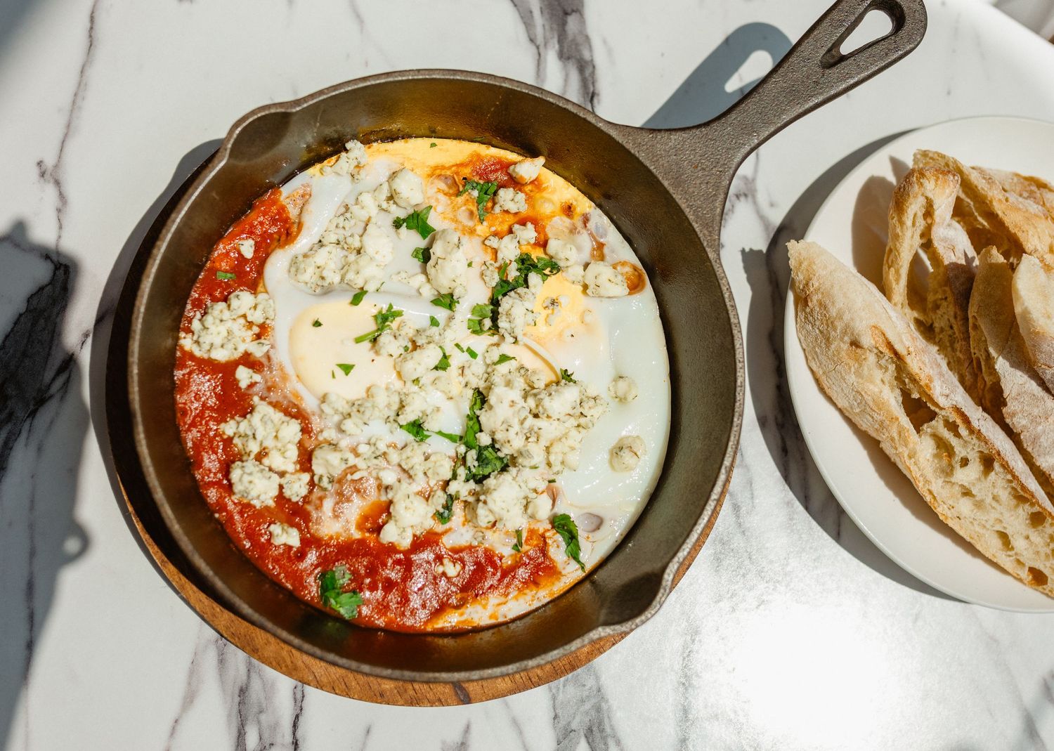 Cast iron skillet of shakshuka topped with feta and fresh herbs, served with sliced rustic bread.