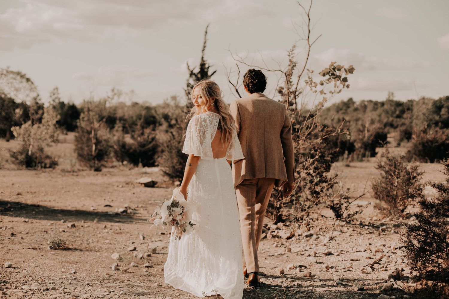 Bride in a backless white dress holding a bouquet walks with groom in a tan suit across a dry desert landscape.