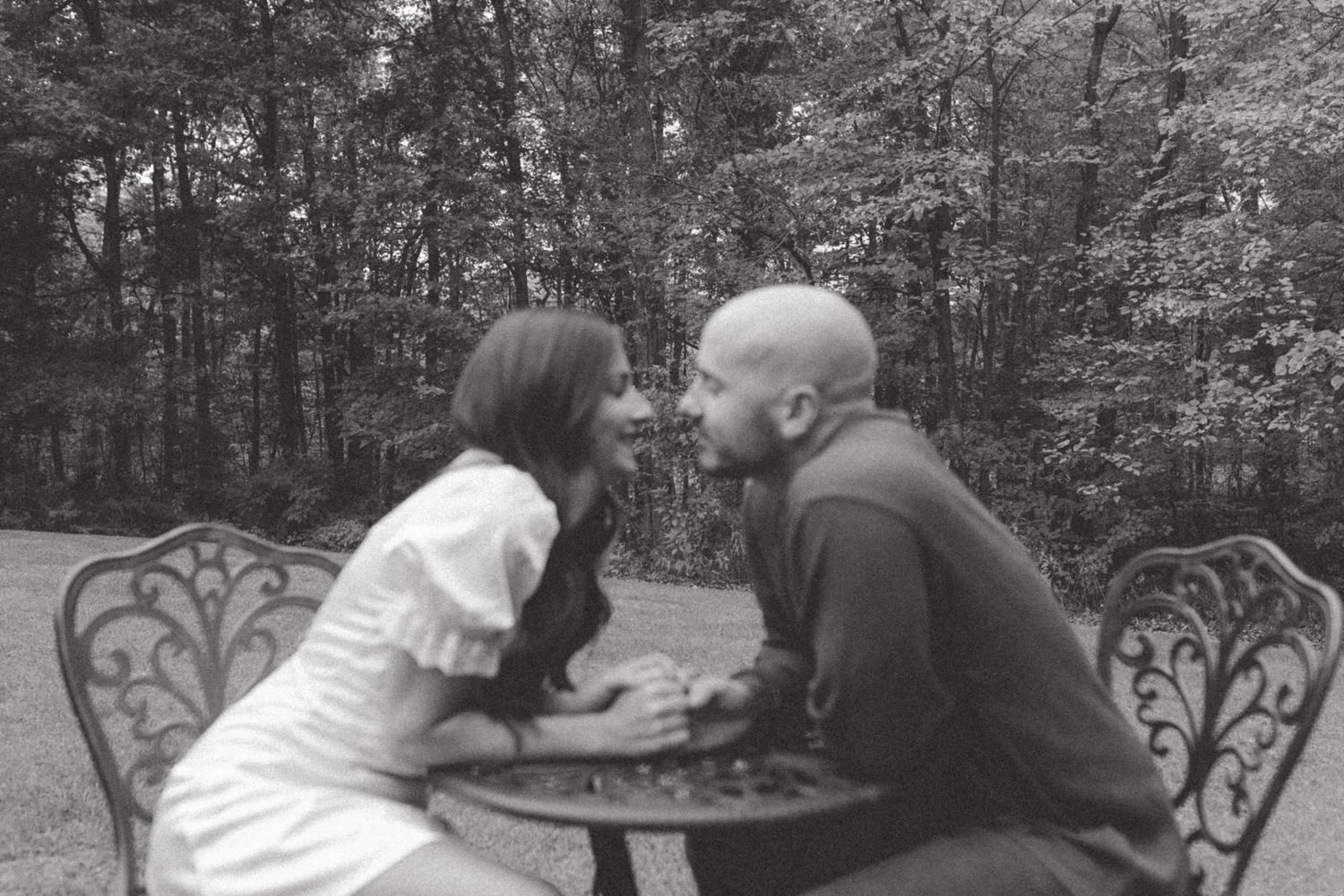 Black and white photo of a couple leaning in close over a vintage metal bistro table with a forest in the background.