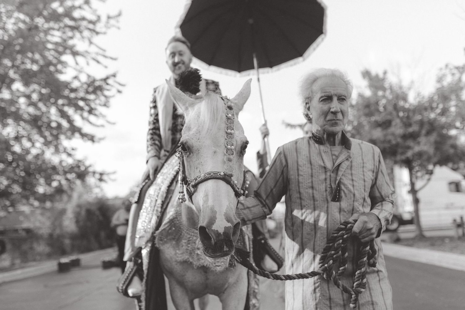 Black and white photo of an older man leading a decorated white horse carrying a rider under a large umbrella along a tree-lined street.