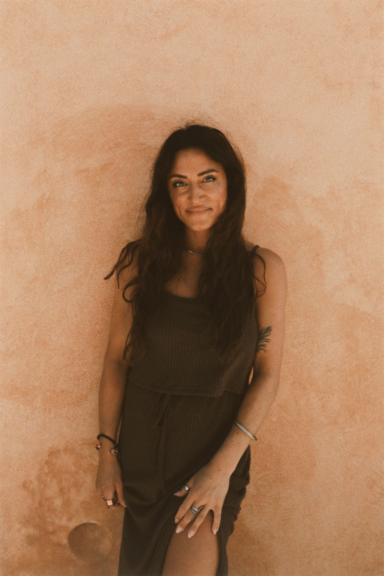 A smiling woman with long wavy dark hair and a black dress poses against a warm terracotta-colored wall.