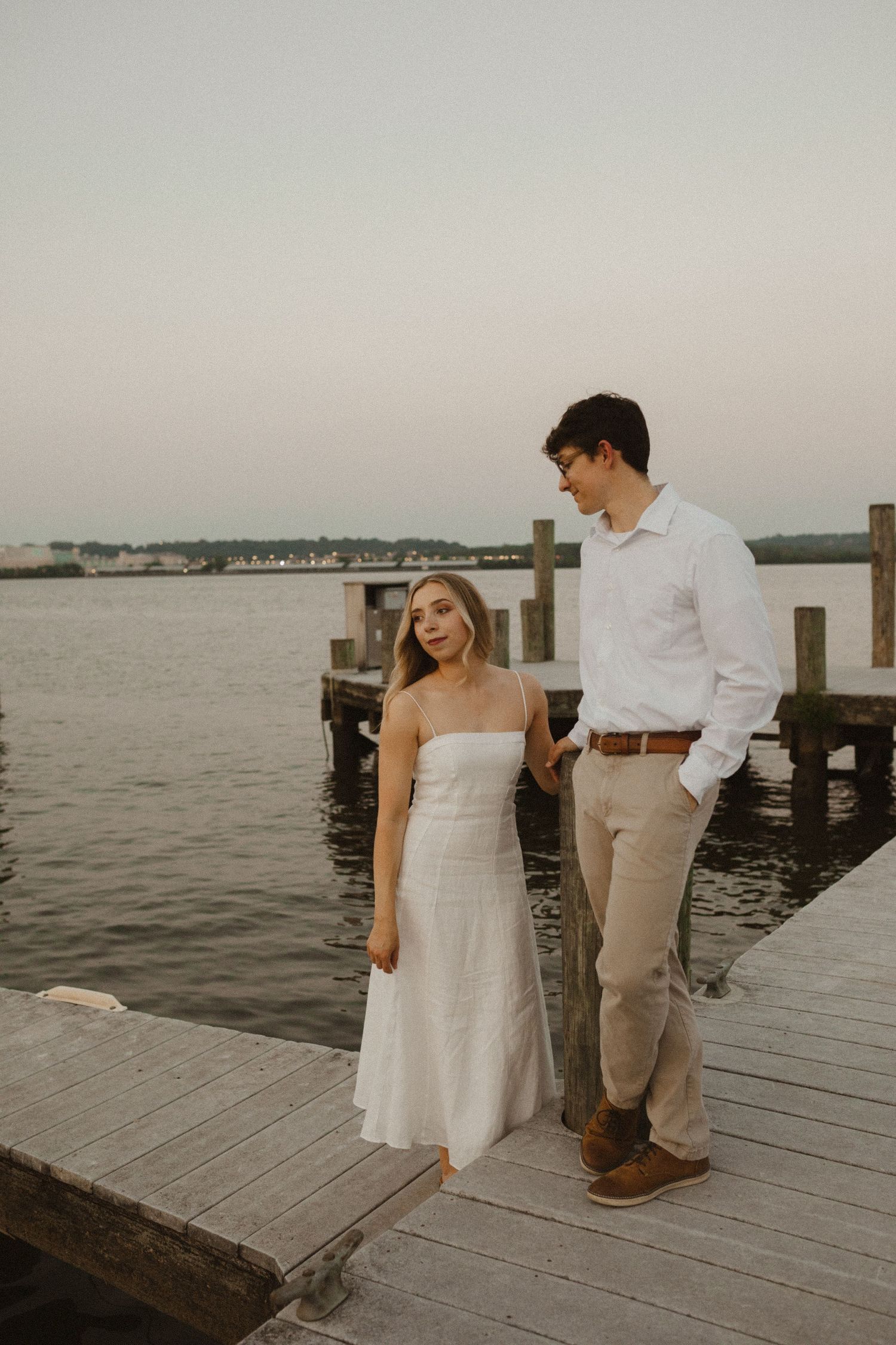 Couple in white dress and button-up shirt pose together on a waterfront wooden pier during an engagement session.