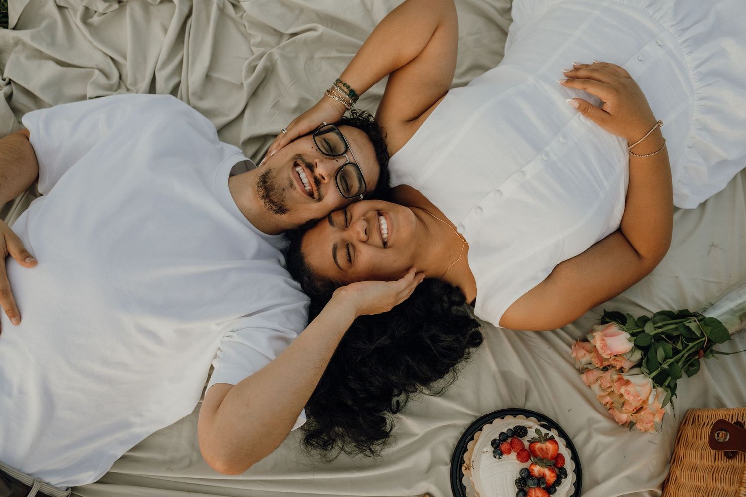 A happy couple lies on a picnic blanket smiling at the camera, with roses and a fruit plate nearby.
