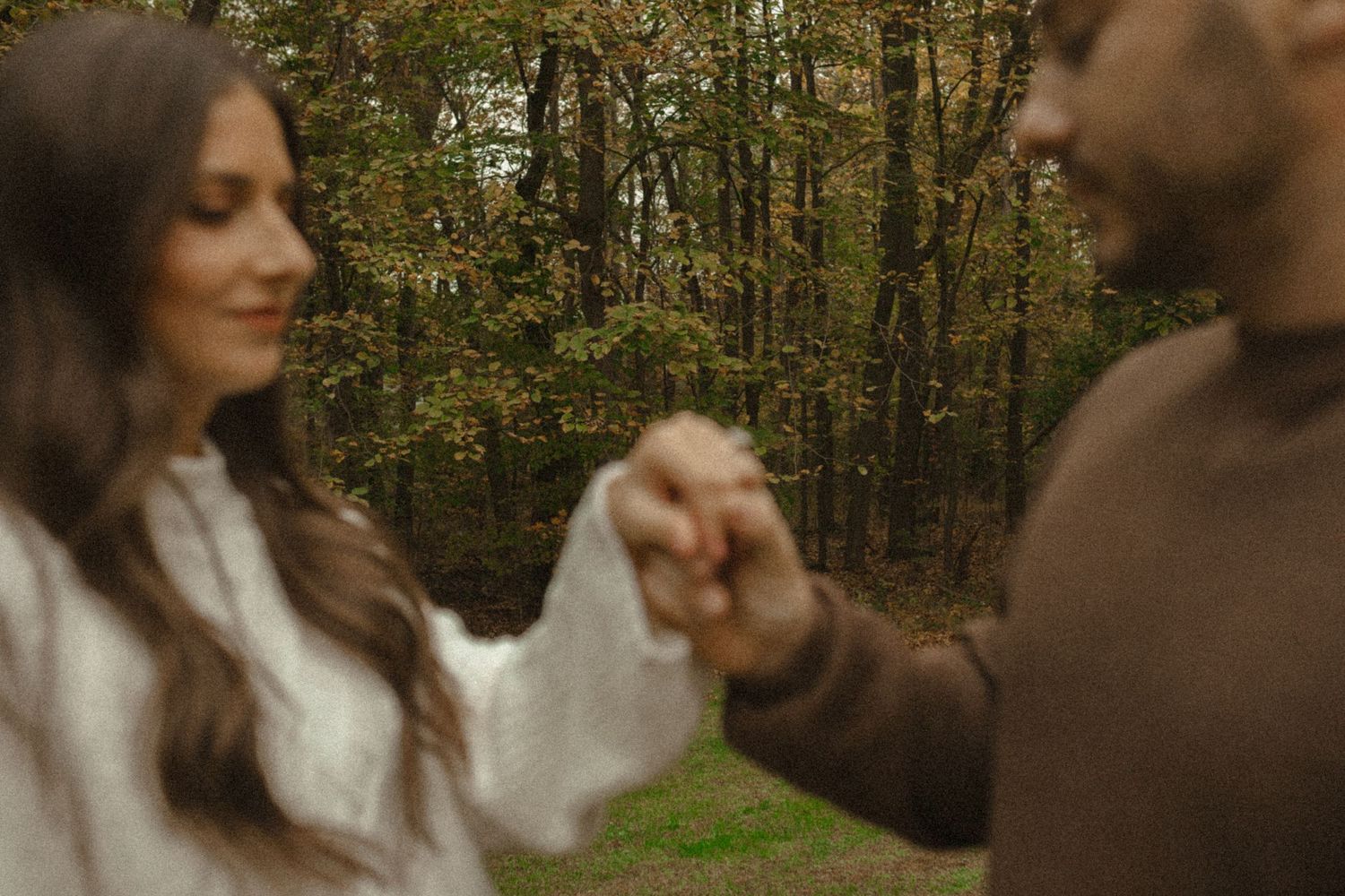Close-up of a smiling woman in a white blouse holding hands with her partner in a softly blurred fall forest.