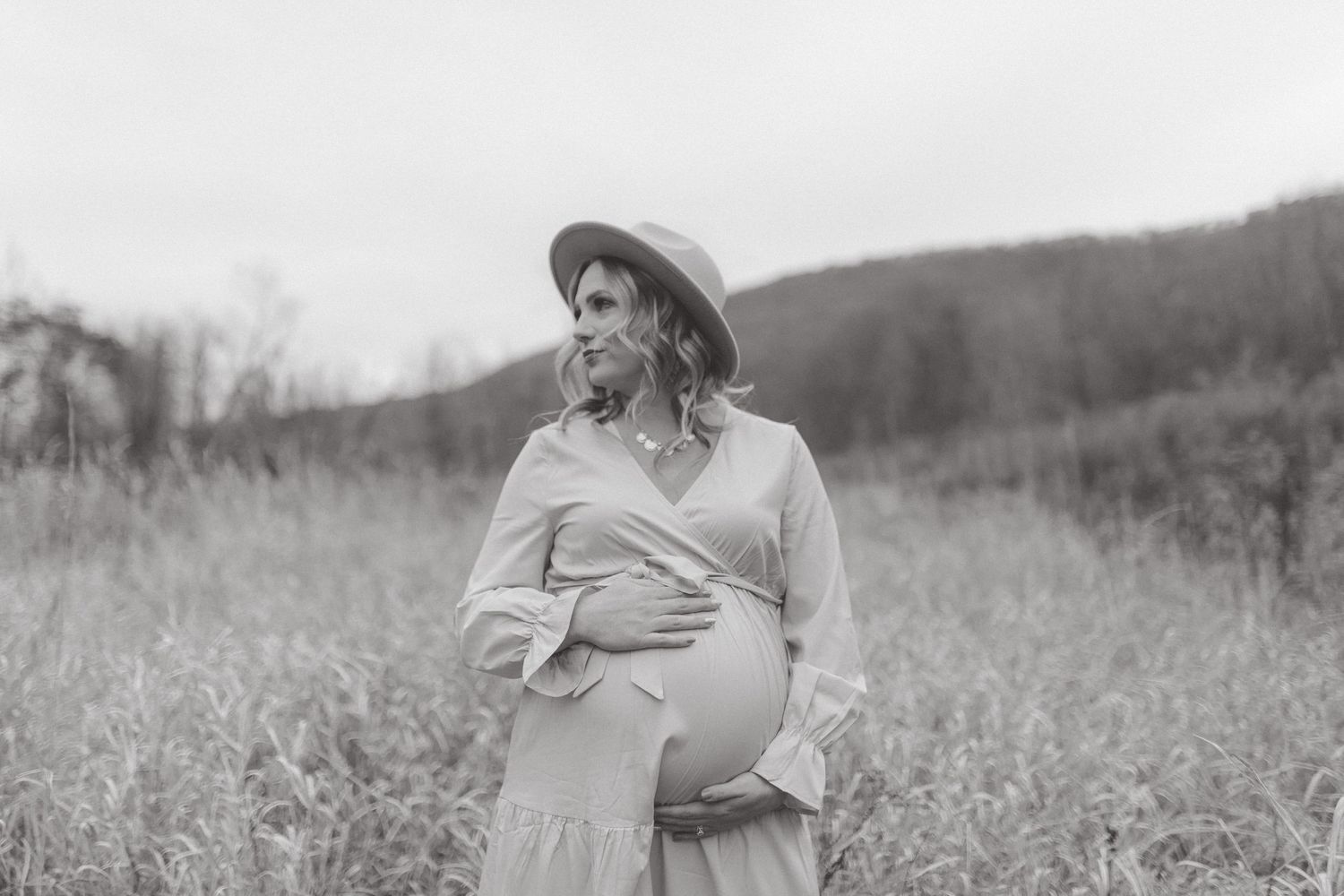 Black and white maternity portrait of a pregnant woman in a hat cradling her belly in a tall grass field.