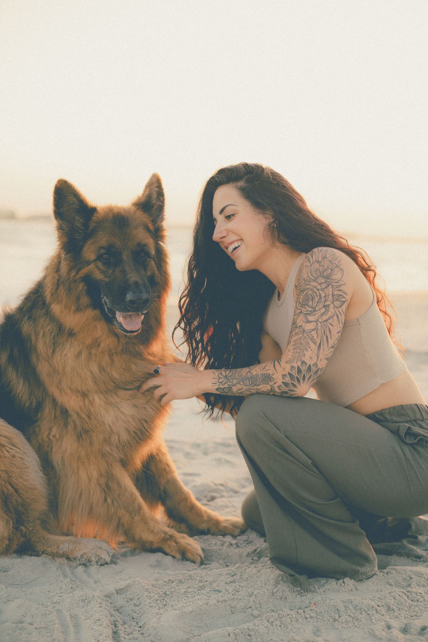 Smiling tattooed woman crouches on the beach and hugs her happy German Shepherd dog in warm golden light.
