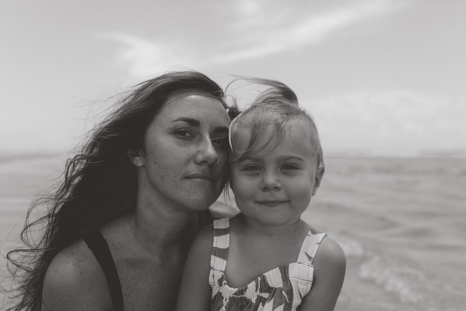 Black and white beach portrait of a mother with long wind-blown hair holding her smiling toddler daughter by the ocean.