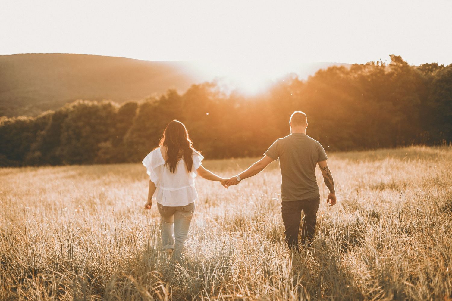 A couple holds hands and walks through a golden meadow toward a glowing sunset with forested hills in the distance.