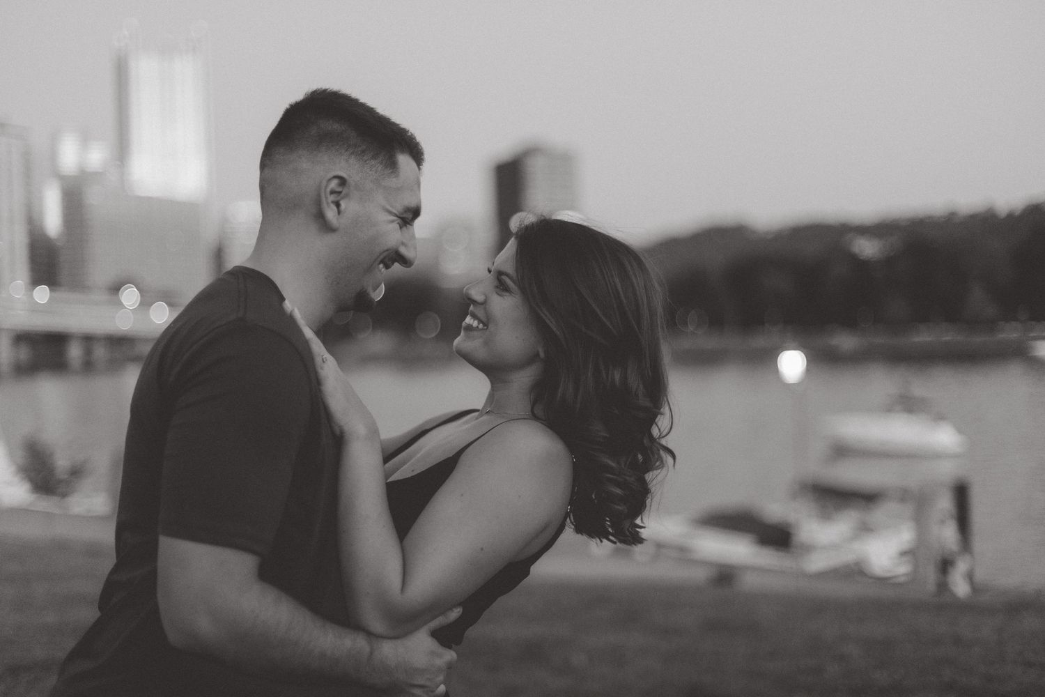 Smiling couple embracing in a black and white engagement photo by a waterfront with city lights in the background.