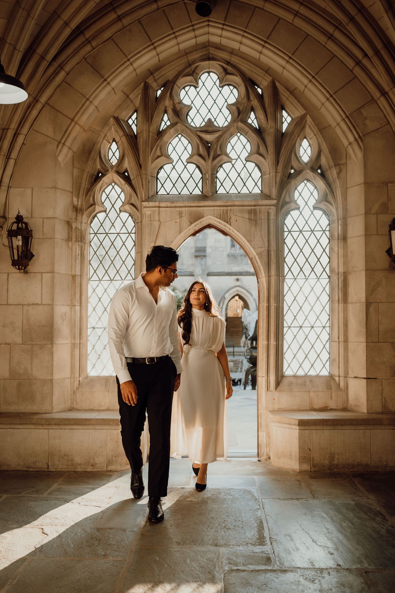 Couple stroll through a sunlit Gothic stone archway with leaded glass windows in a romantic engagement photo.