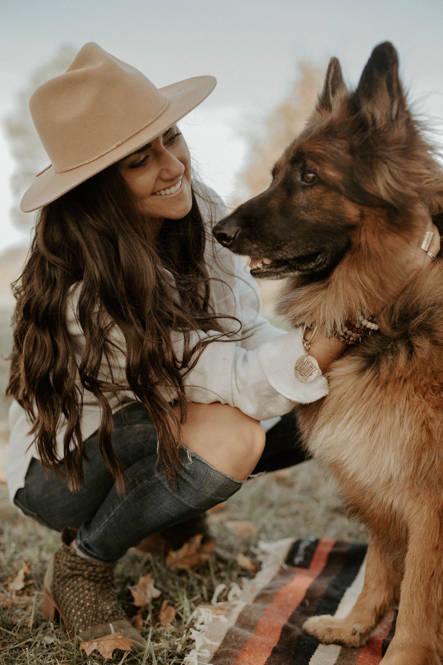 Smiling woman in a tan felt hat crouching beside a German Shepherd wearing a pendant collar on a striped blanket in autumn.