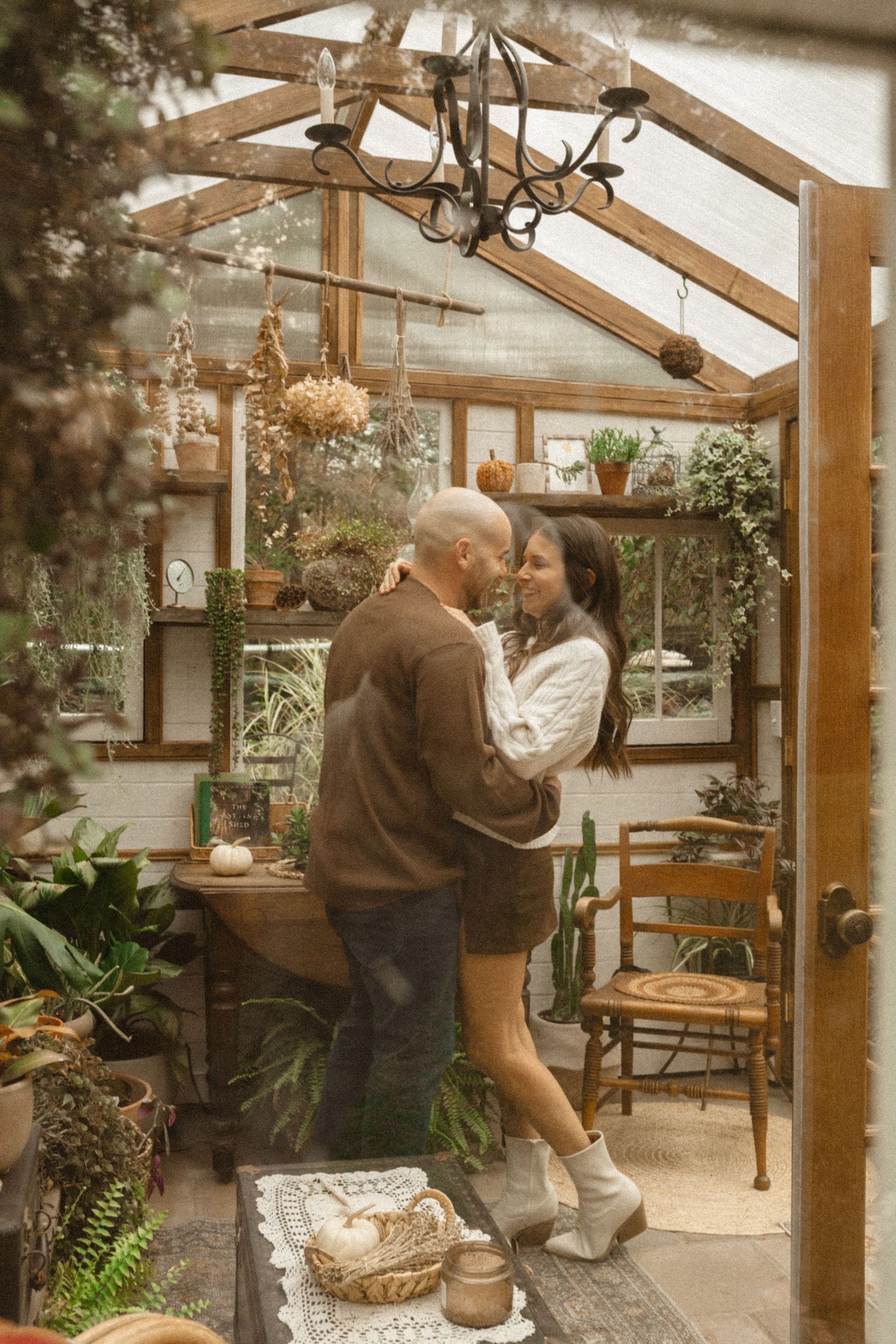 Couple embraces and smiles inside a charming greenhouse filled with hanging plants, a wooden chair, and a chandelier.