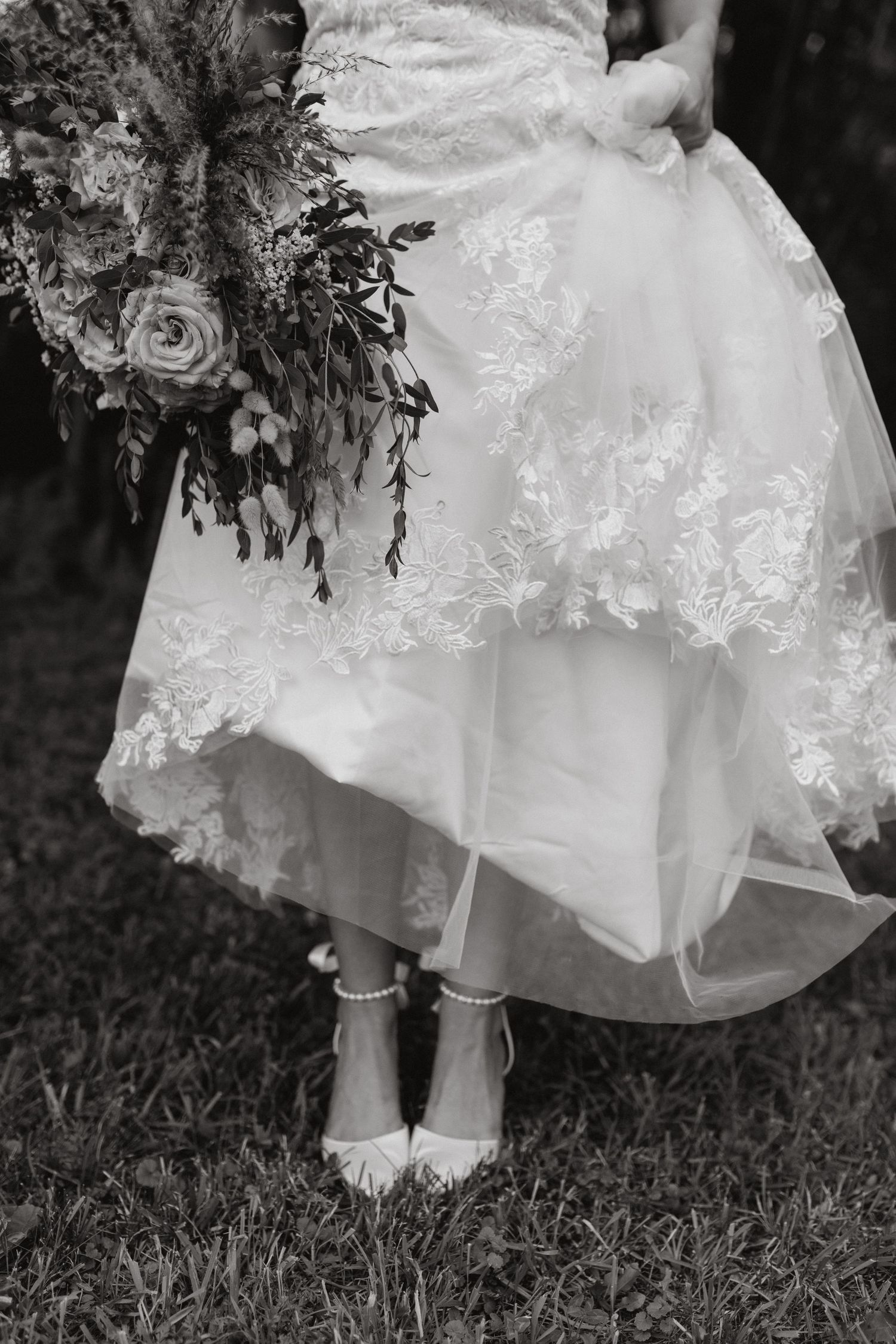 Black and white photo of a bride in a lace tulle wedding gown and beaded flat shoes holding a lush floral bouquet on grass.