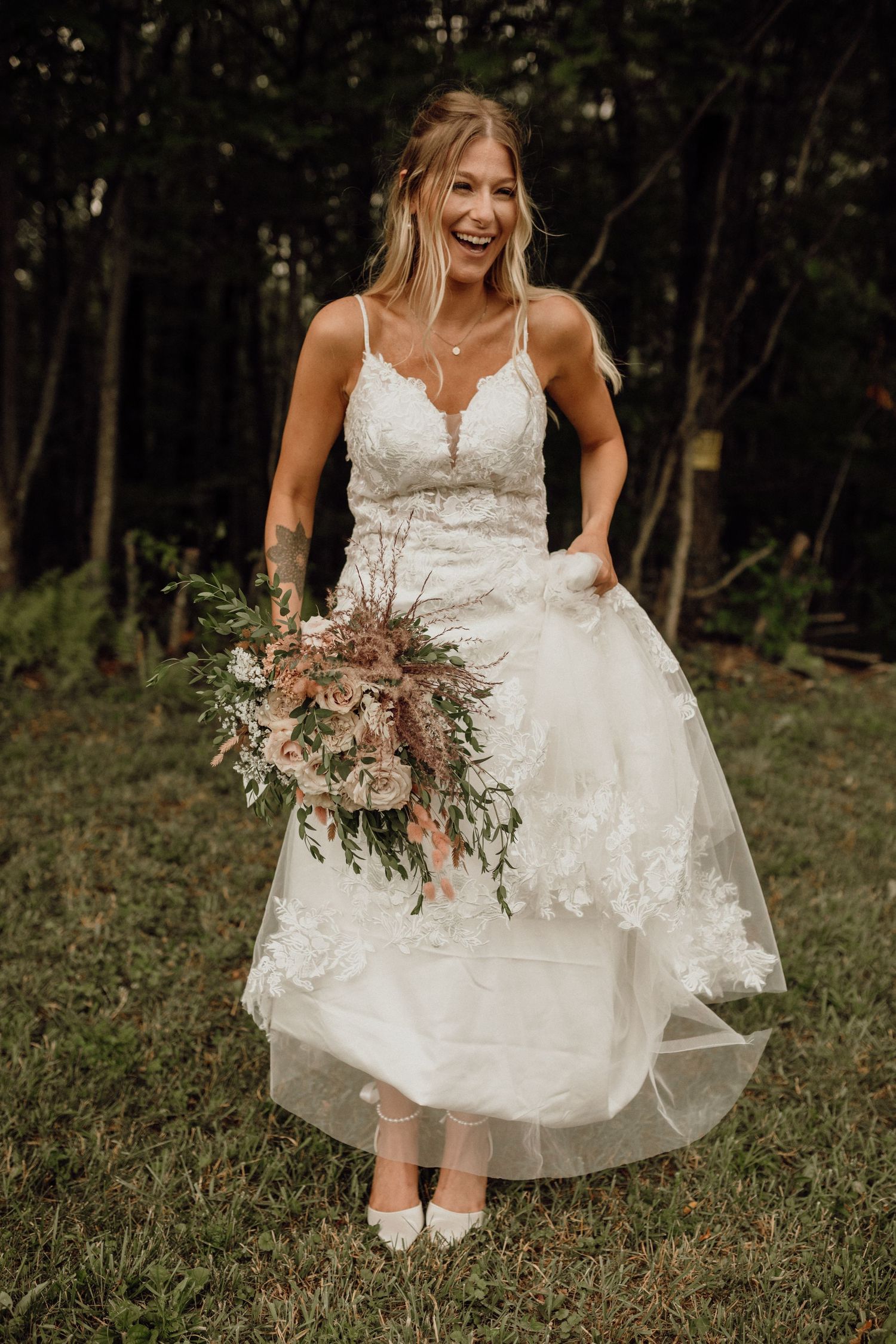 Smiling bride in a lace wedding dress holding a rustic floral bouquet while standing on grass near the woods.
