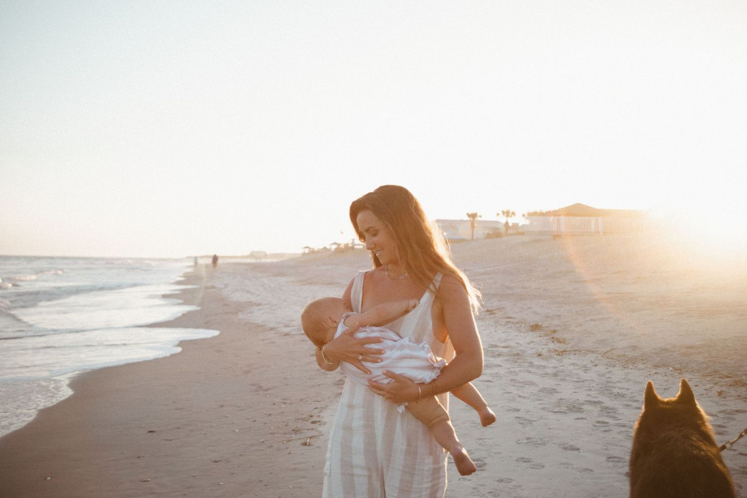 Mother in a striped dress cradling her baby on a golden sunset beach with a dog in the foreground.