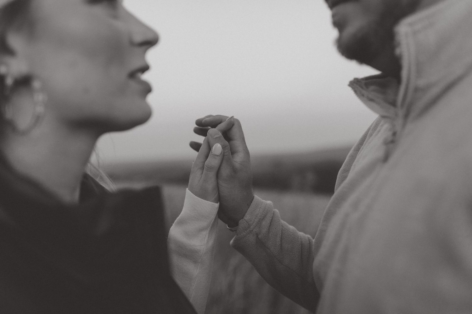 Black and white close-up of a couple facing each other outdoors, their fingers softly intertwined between them.