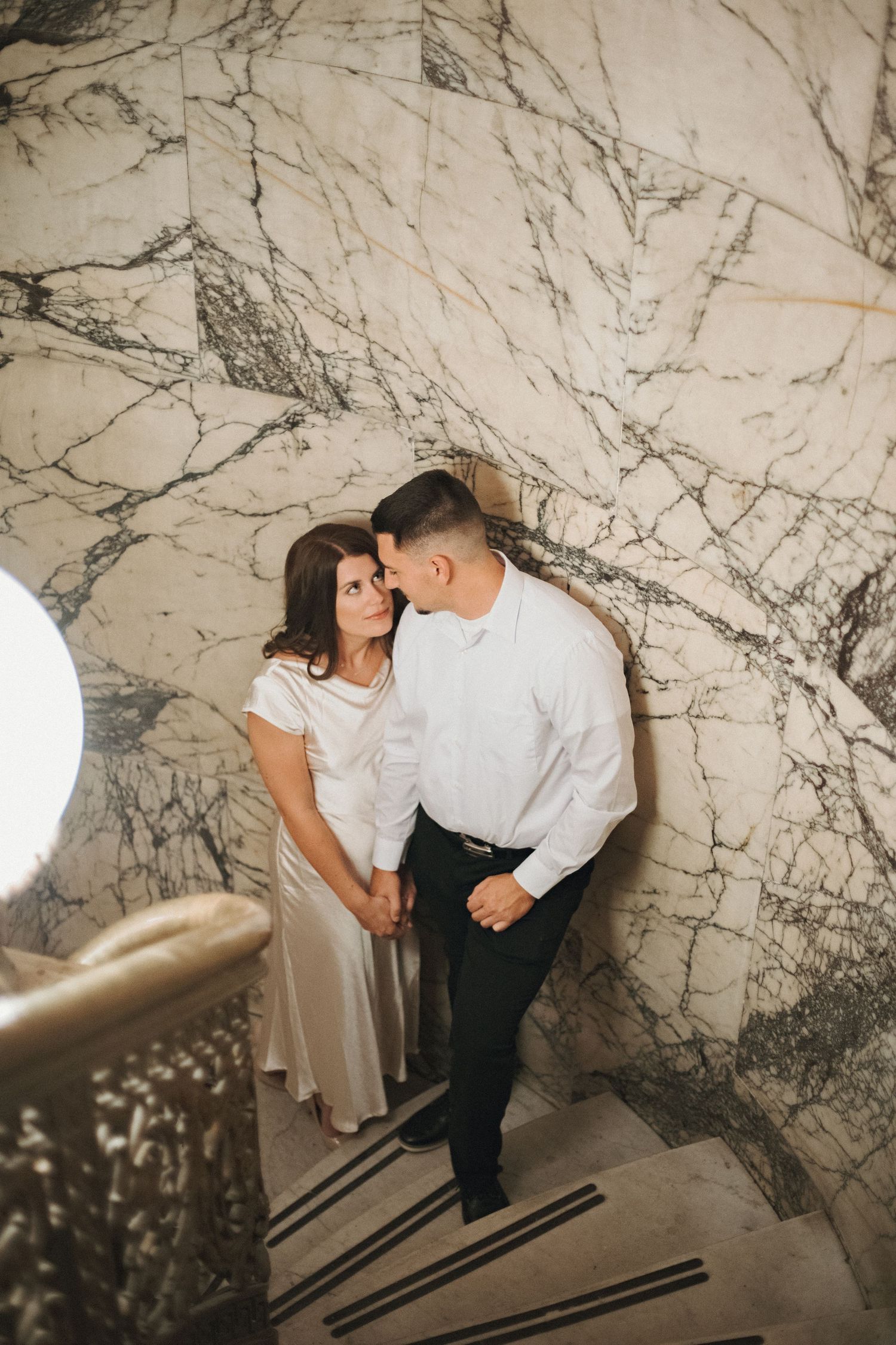 Couple holding hands and gazing at each other on a marble staircase in a historic building during an engagement photo session.