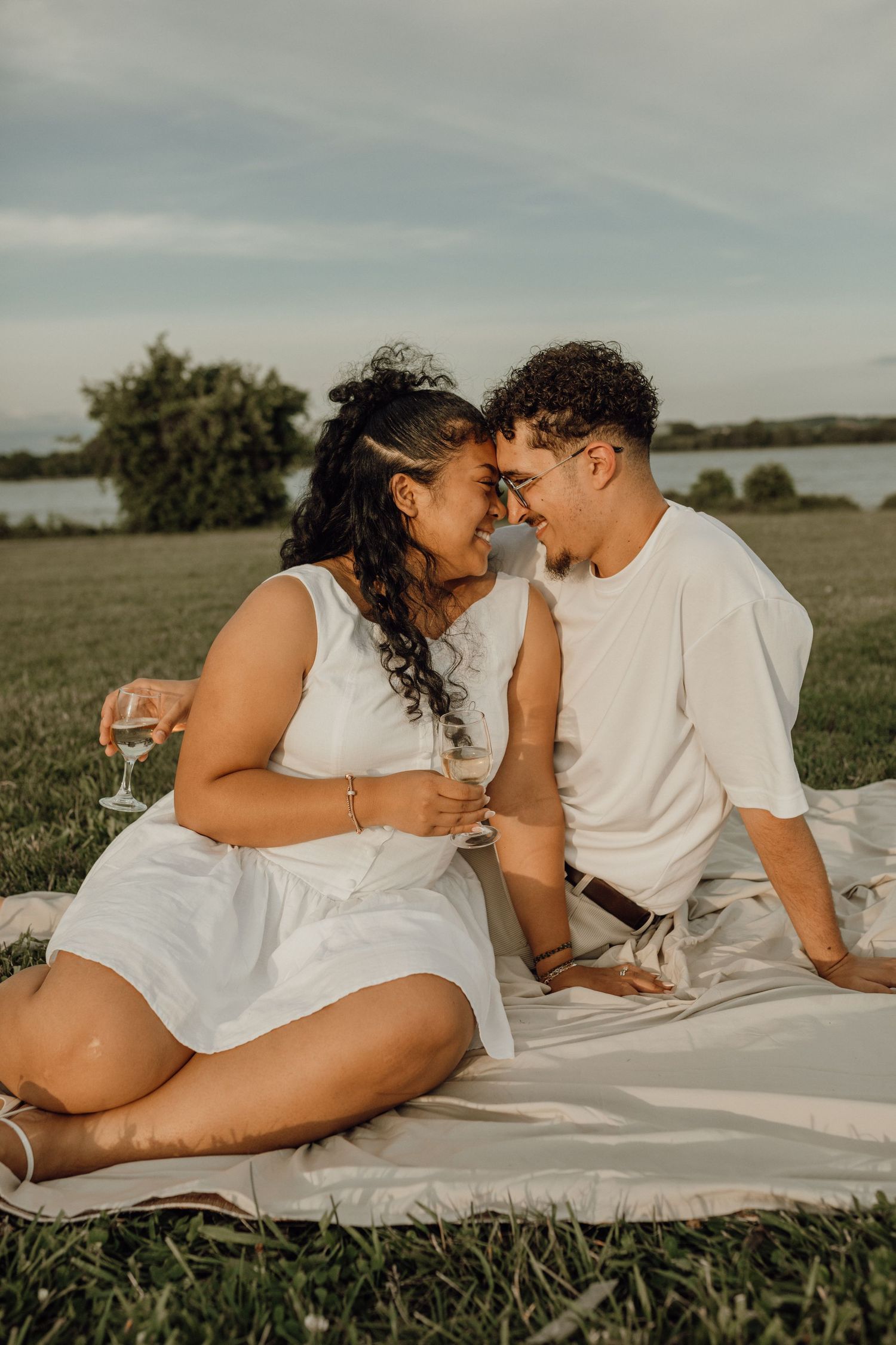 A smiling couple in white outfits touch foreheads while holding wine glasses on a picnic blanket by a lake.