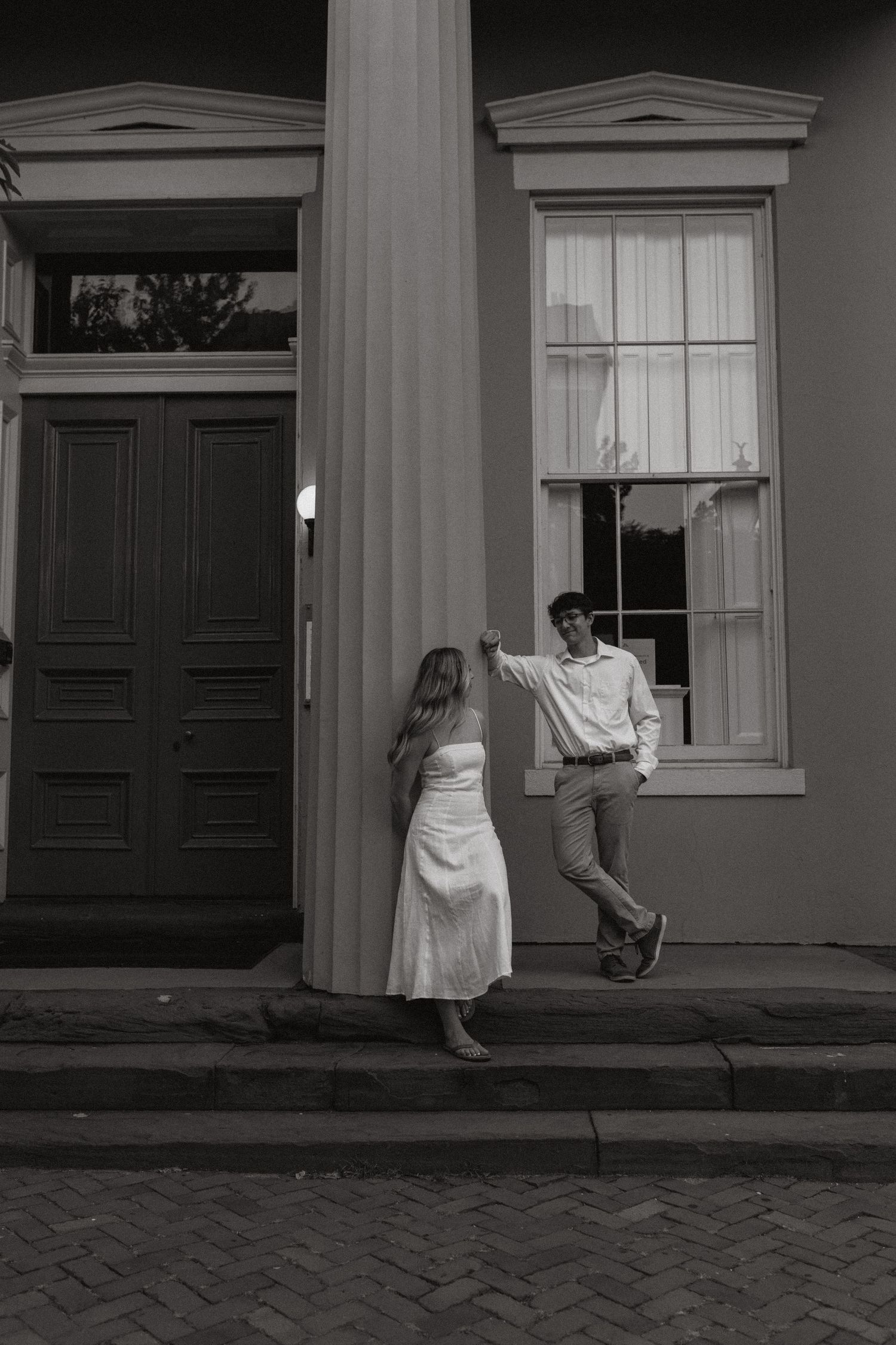 Black and white photo of a man leaning on a column while a woman in a white dress stands beside a historic building.