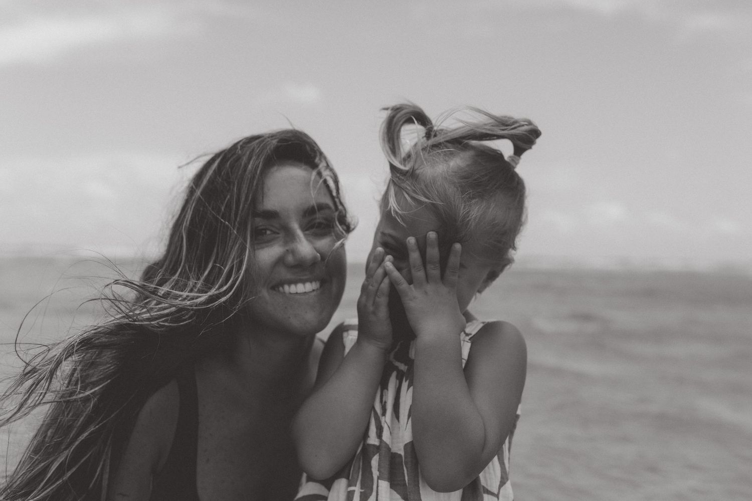 Black and white photo of a smiling woman with windswept hair holding a little girl on a beach with the ocean behind.