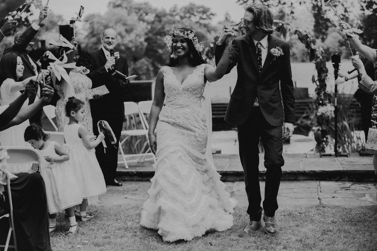Smiling bride and groom walk hand in hand down the aisle as guests cheer and toss petals after their outdoor wedding ceremony.