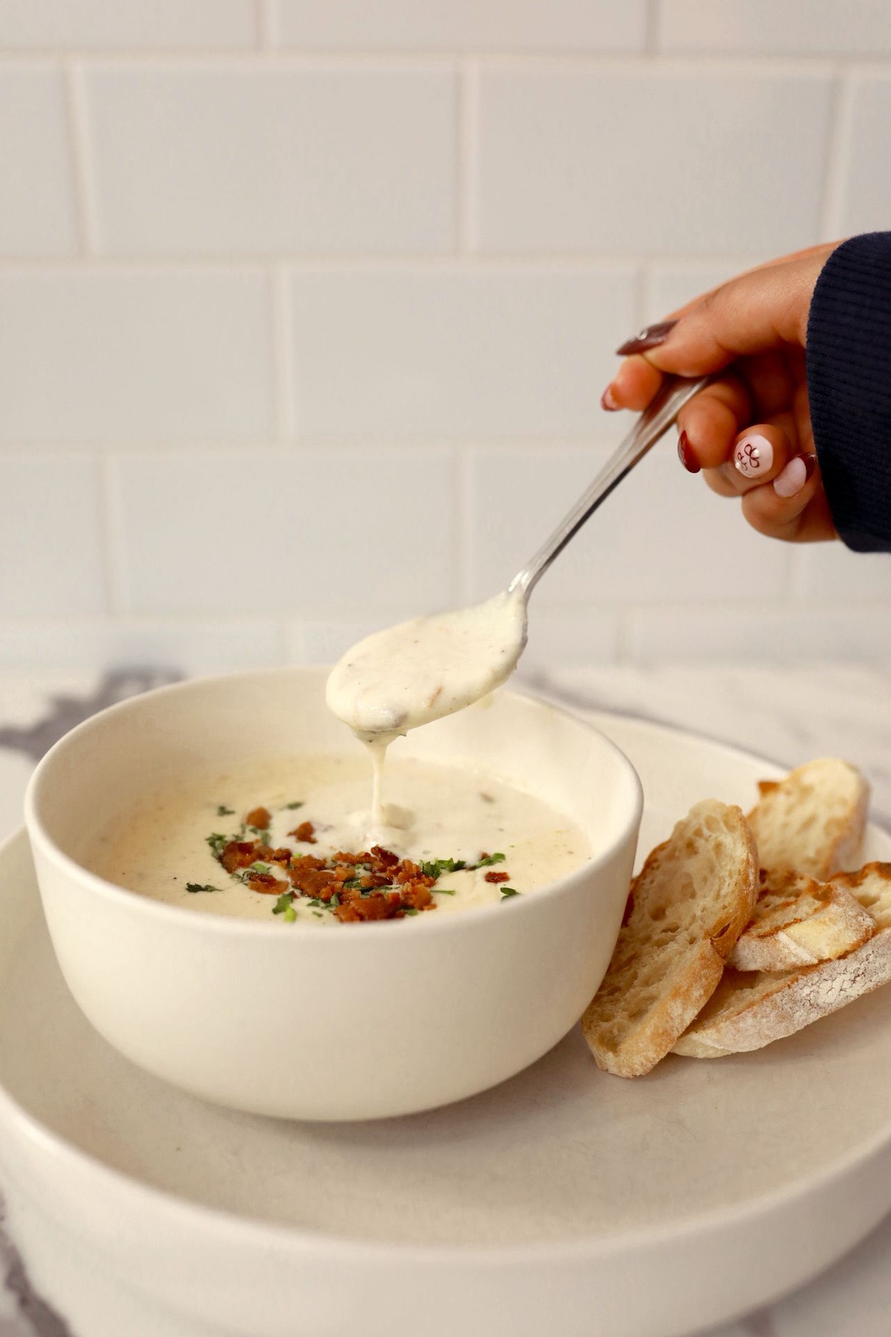 Hand lifting a spoonful of creamy soup from a white bowl topped with bacon and herbs, with sliced toasted bread on the plate.