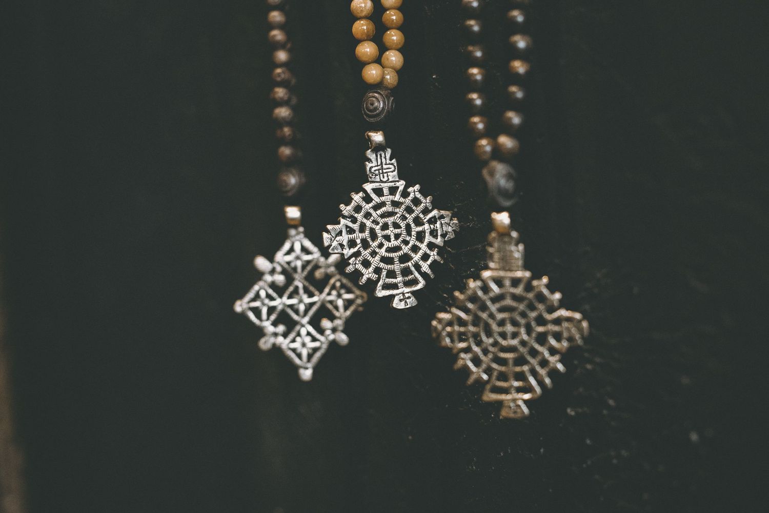 Three ornate silver cross pendants hanging from wooden bead necklaces against a dark background.
