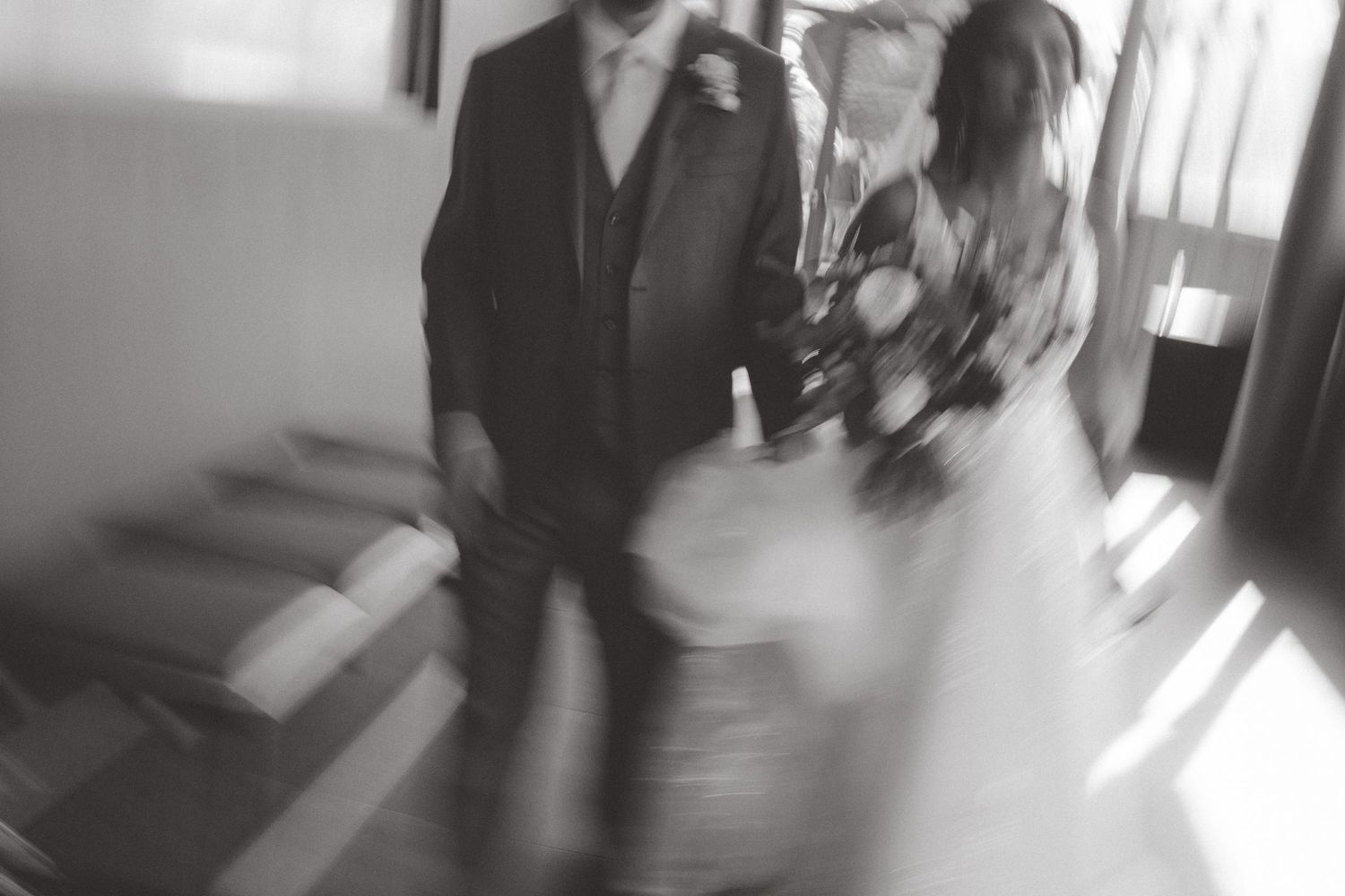 Blurred black and white photo of a bride and groom in motion, the bride holding a bouquet, with soft light streaming in.