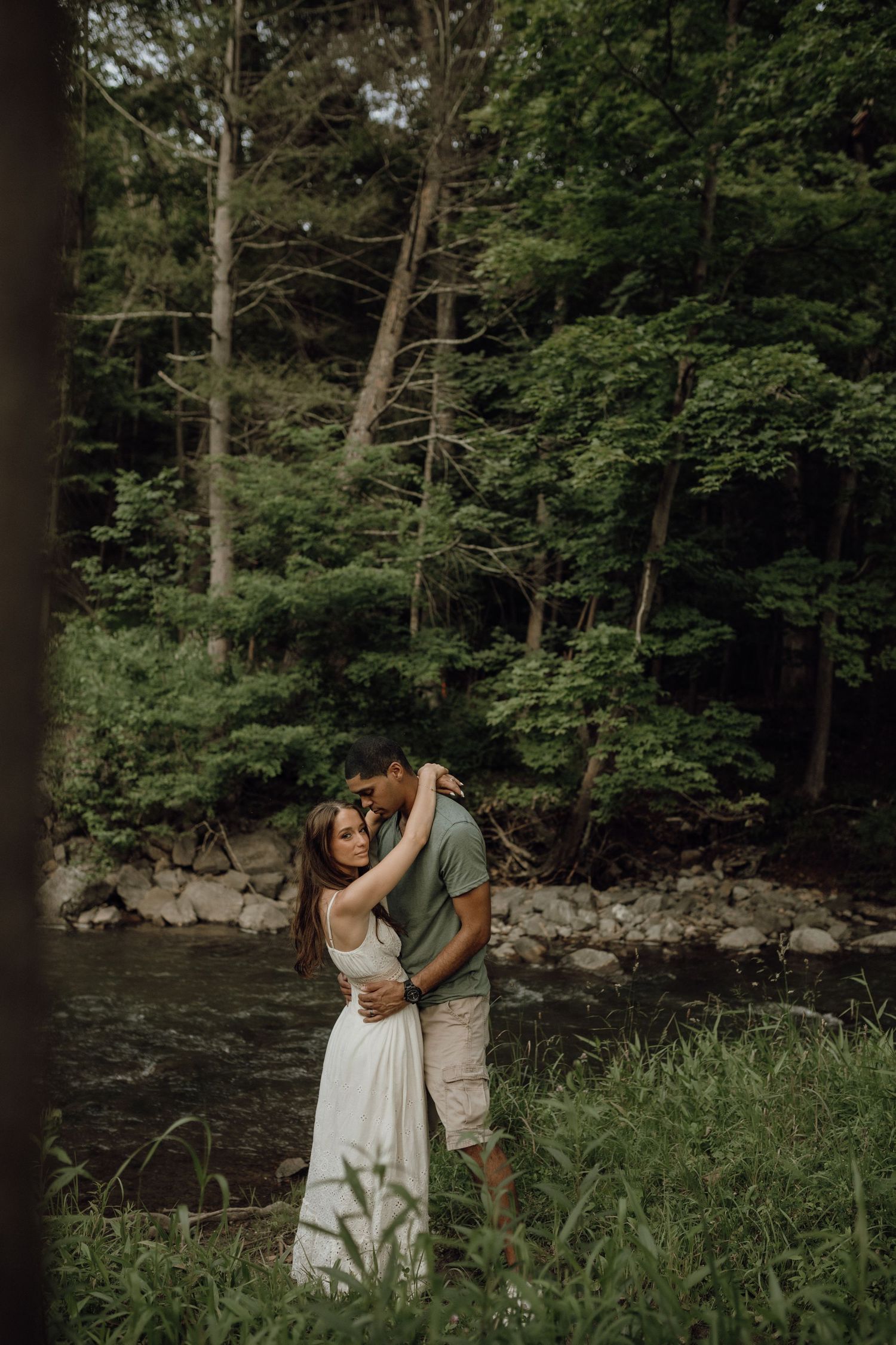 A couple embraces beside a rocky forest stream surrounded by tall trees and lush green grass.