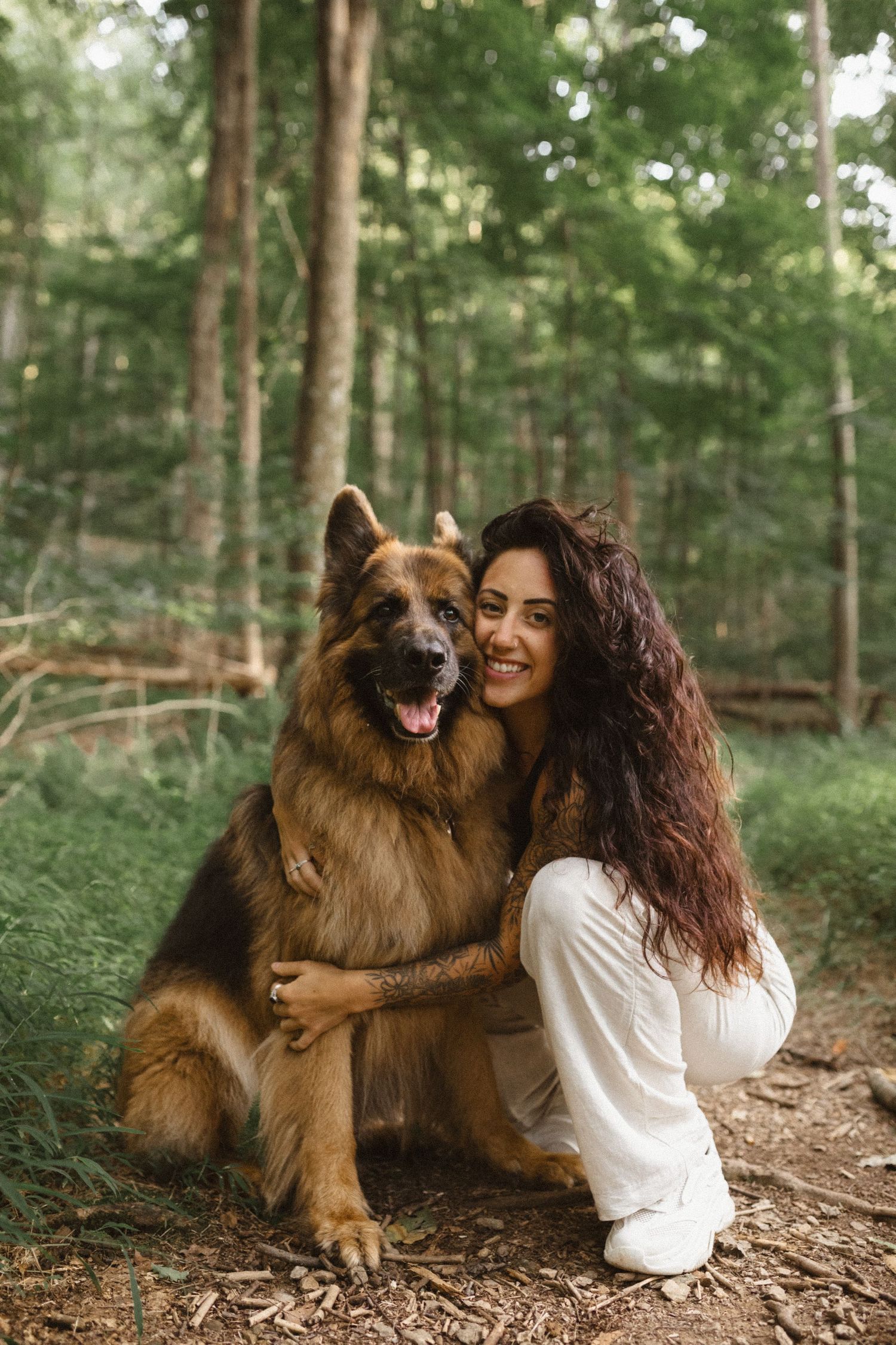 Woman in white pants kneels in a sunlit forest and hugs her German Shepherd dog with its tongue out.