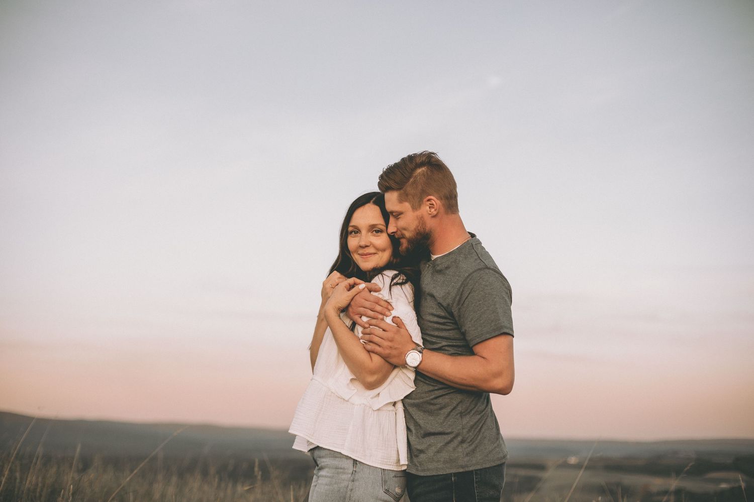 Smiling woman in a white blouse embraced from behind by her bearded partner during a sunset engagement session outdoors.