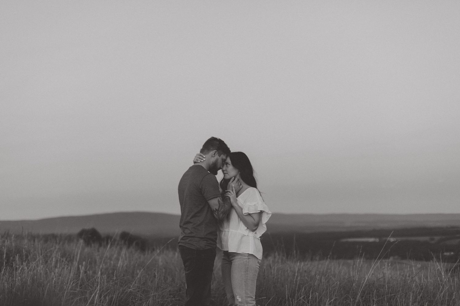 Black and white portrait of a couple embracing forehead to forehead in a grassy field at dusk with distant hills.
