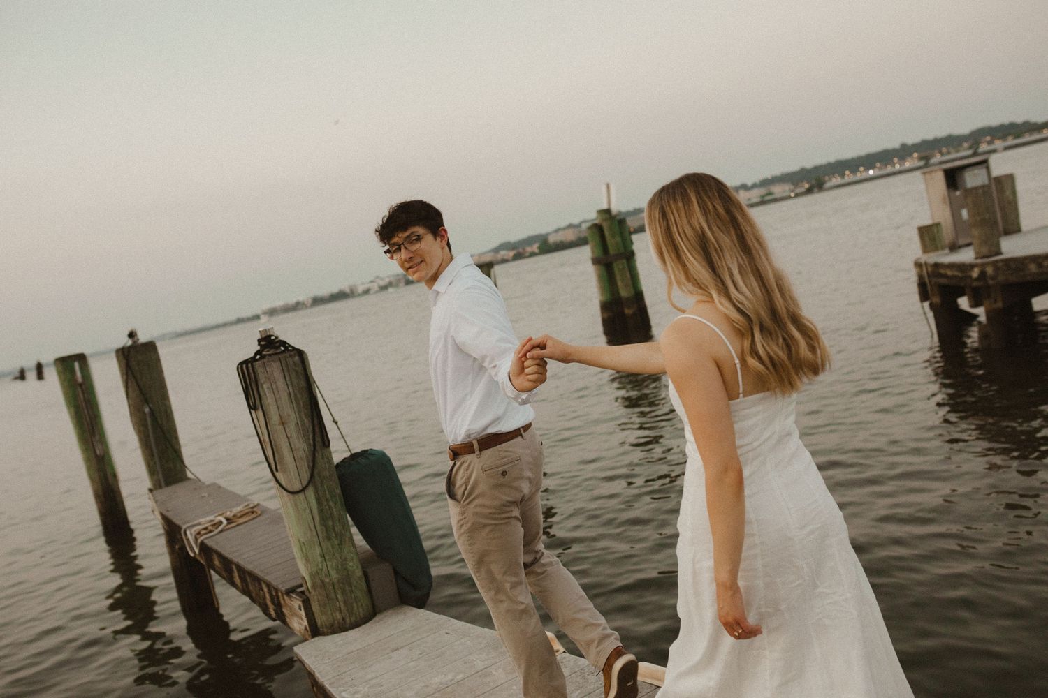 Woman in a white dress holds a man's hand as they walk along a wooden dock by the water during an engagement photo shoot.