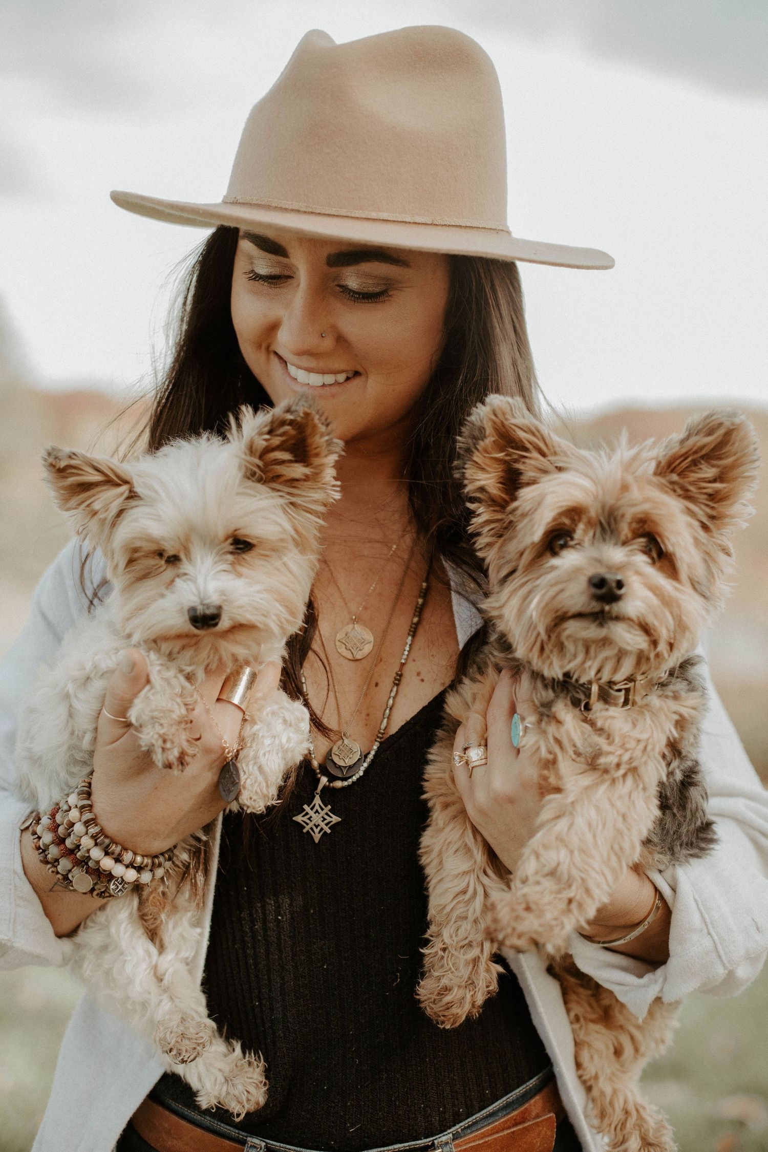 Smiling woman in a felt hat and layered necklaces holding two fluffy Yorkshire terrier dogs in a desert landscape.