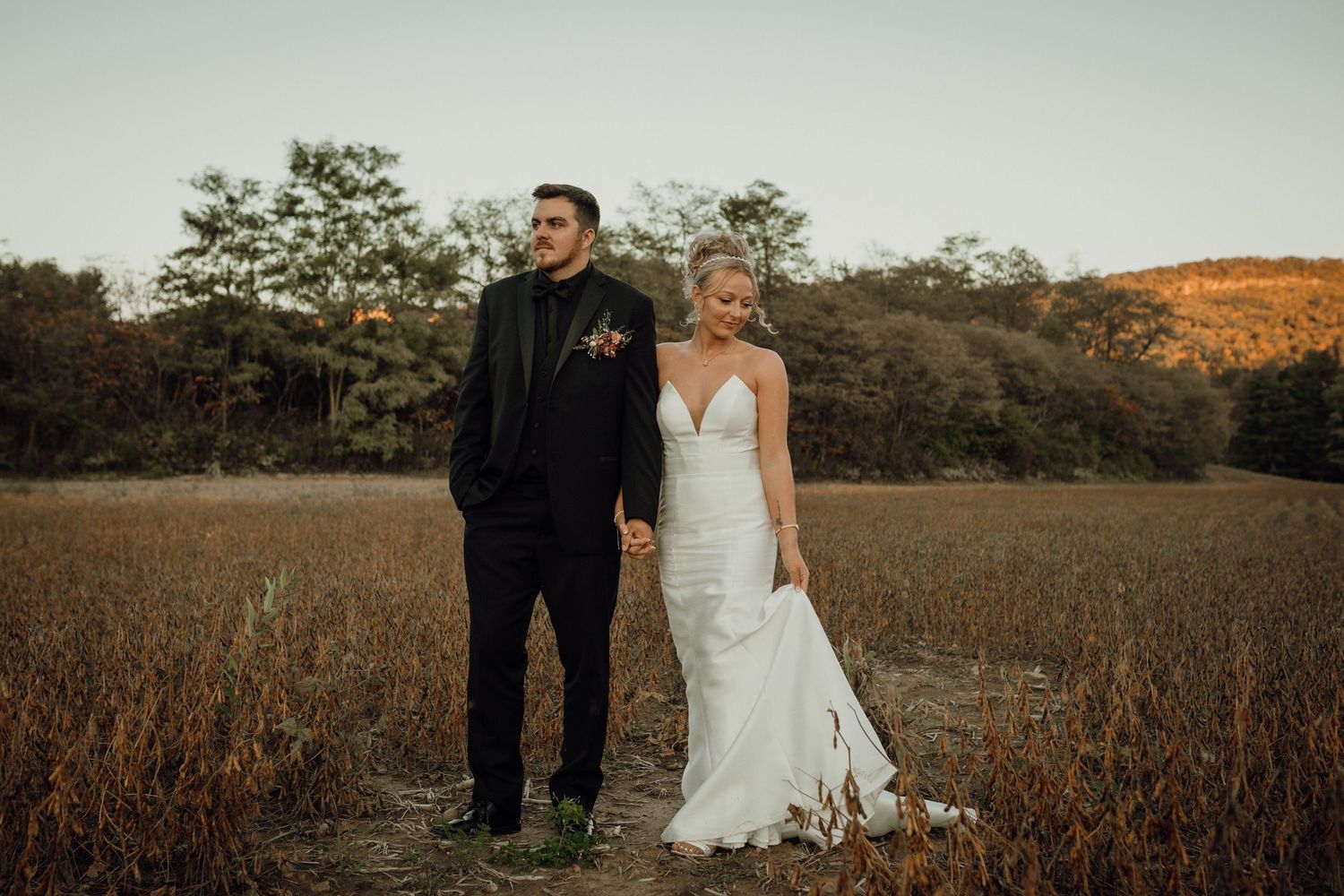 Bride in a white gown and groom in a black suit hold hands in a golden field with trees behind them.