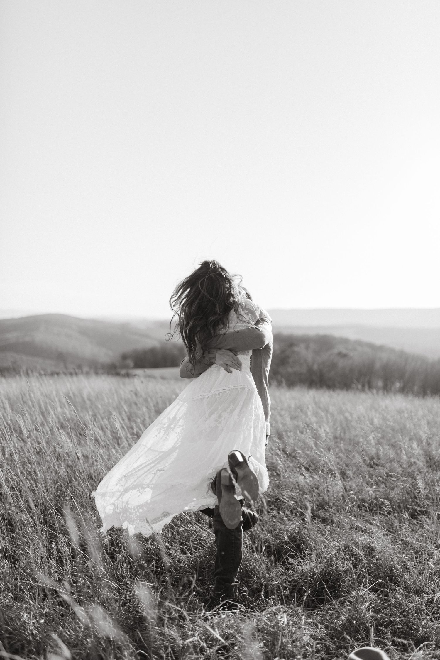 Black and white photo of a bride in a flowing white dress and cowboy boots embracing her partner in a grassy hilltop field.