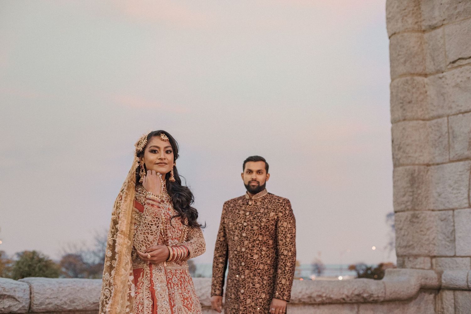 Bride in a traditional embroidered lehenga and groom in a patterned sherwani stand beside a stone pillar at dusk.