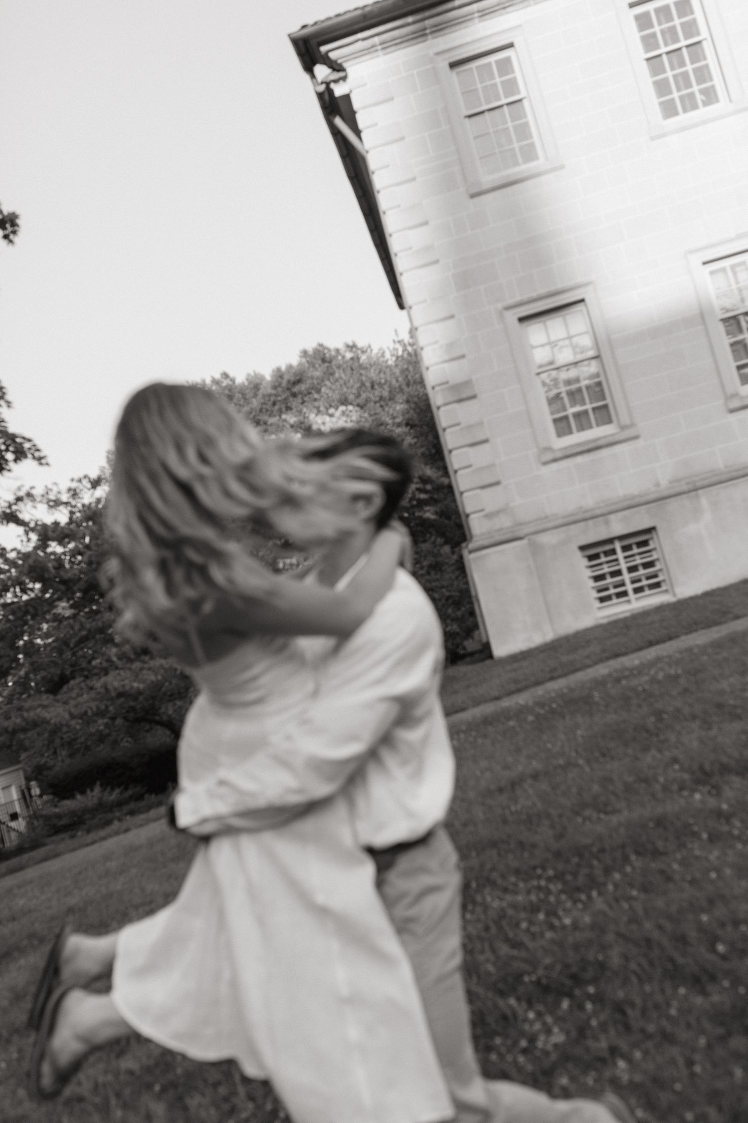 A couple embraces and spins on a lawn beside a historic stone building in a candid black and white engagement photo.