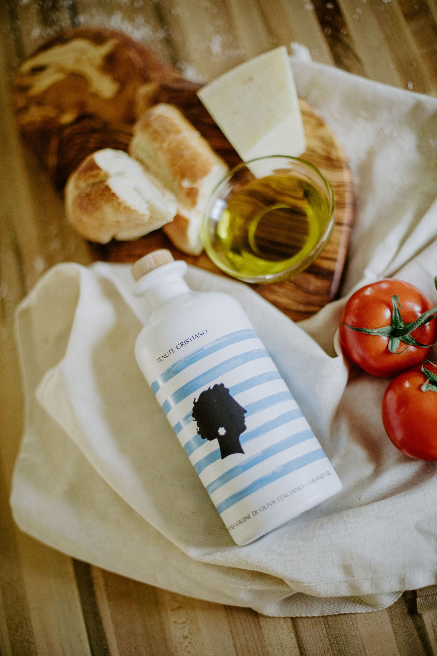 Blue striped bottle of extra virgin olive oil with bread, cheese and ripe tomatoes on a rustic wooden board.
